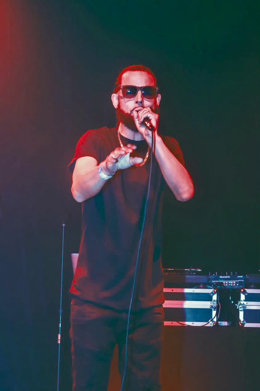 Person with sunglasses singing into a microphone on stage, with DJ equipment in the background.