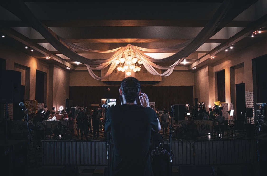 Person with back facing camera watching a stage with a chandelier and draped fabric in a dimly lit event hall.