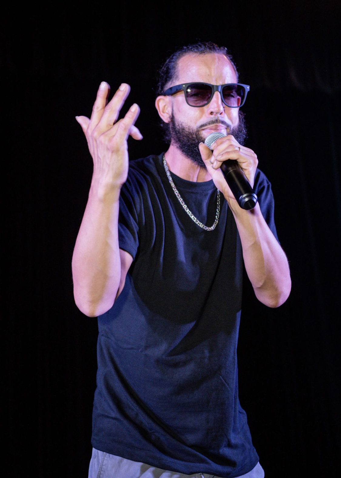 Person with sunglasses and beard singing into a microphone, gesturing with hand, on stage with dark background.