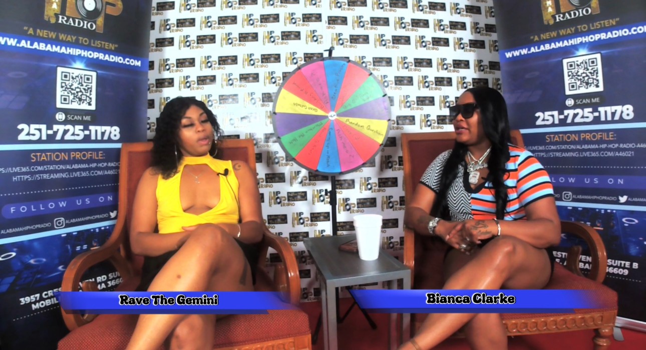 Two women sit on chairs in a studio with a colorful wheel between them, background with radio station banners, names displayed.