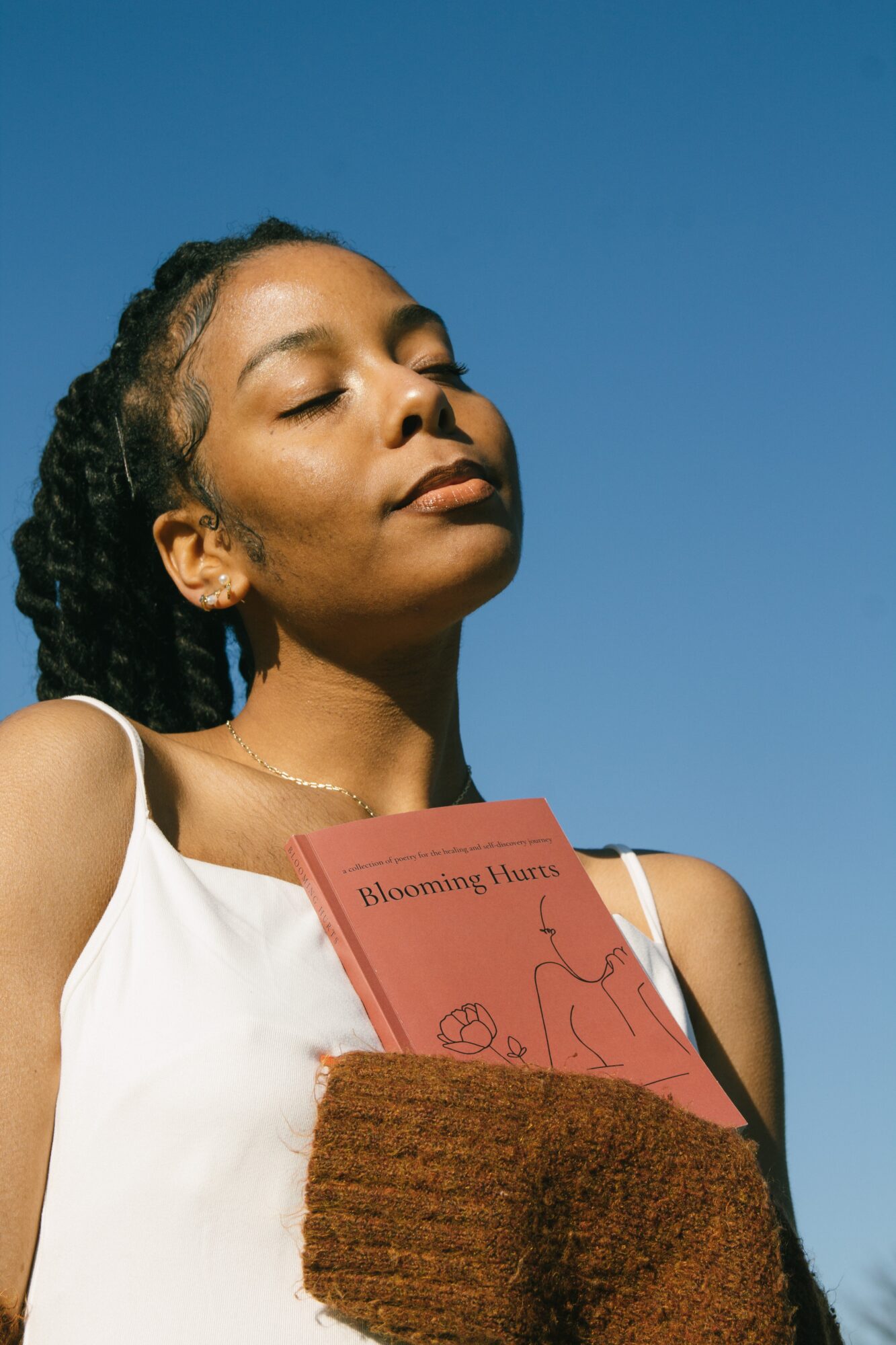Young woman with closed eyes and relaxed expression holding a pink book outdoors against a clear blue sky.