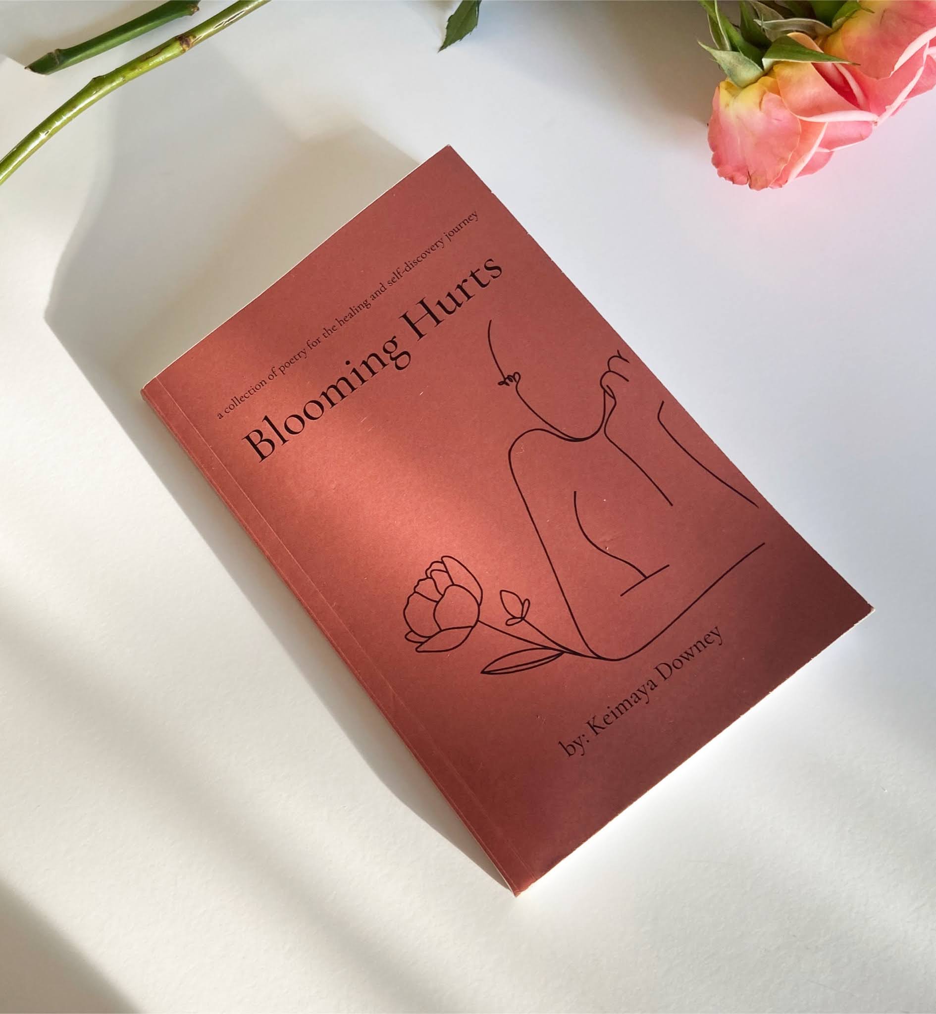 Book titled 'Blooming Hurts' with a line drawing of a person and a flower on a white surface, pink flowers nearby.