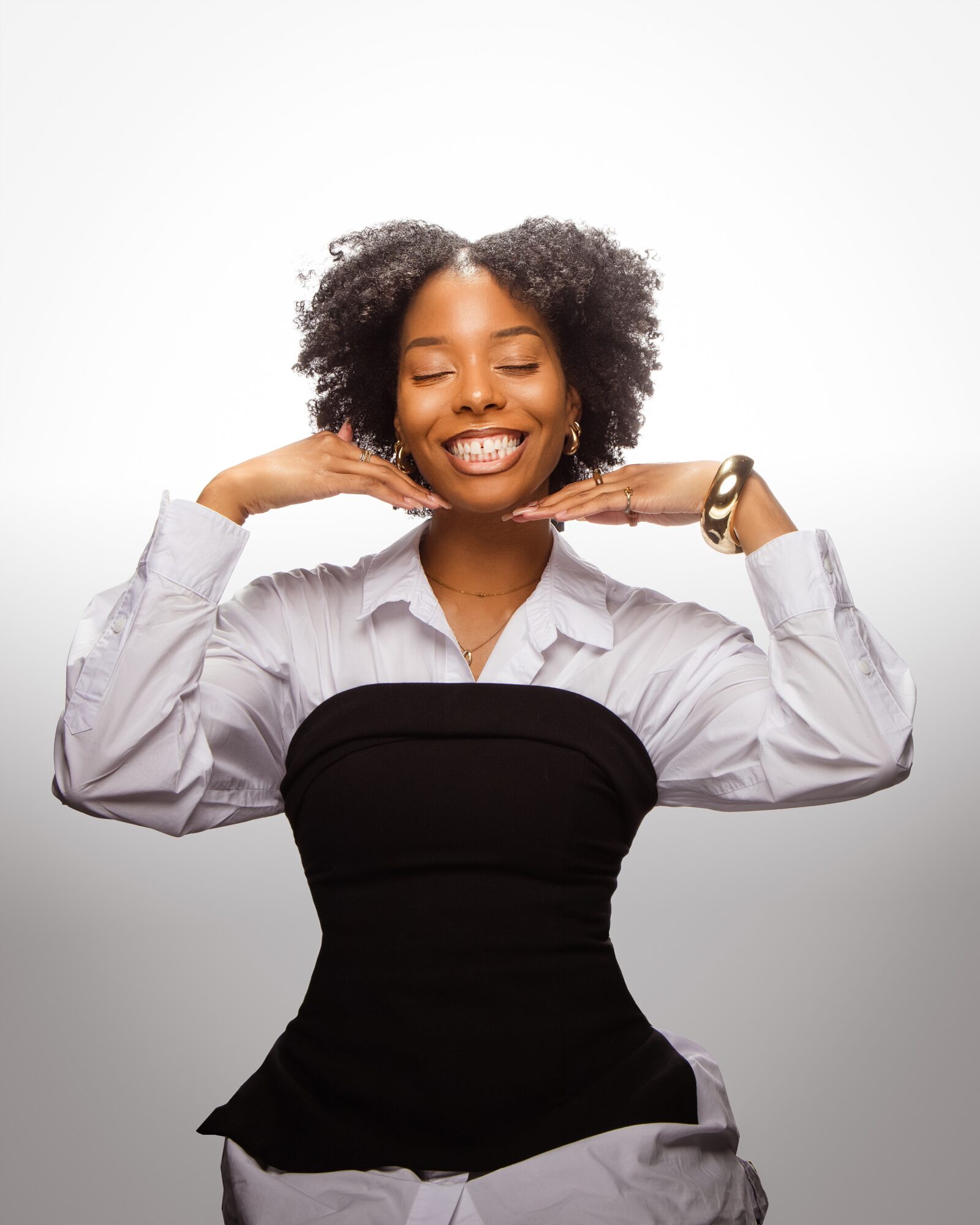 Smiling woman with curly hair, wearing a white shirt and black strapless top, poses with hands near face, eyes closed.
