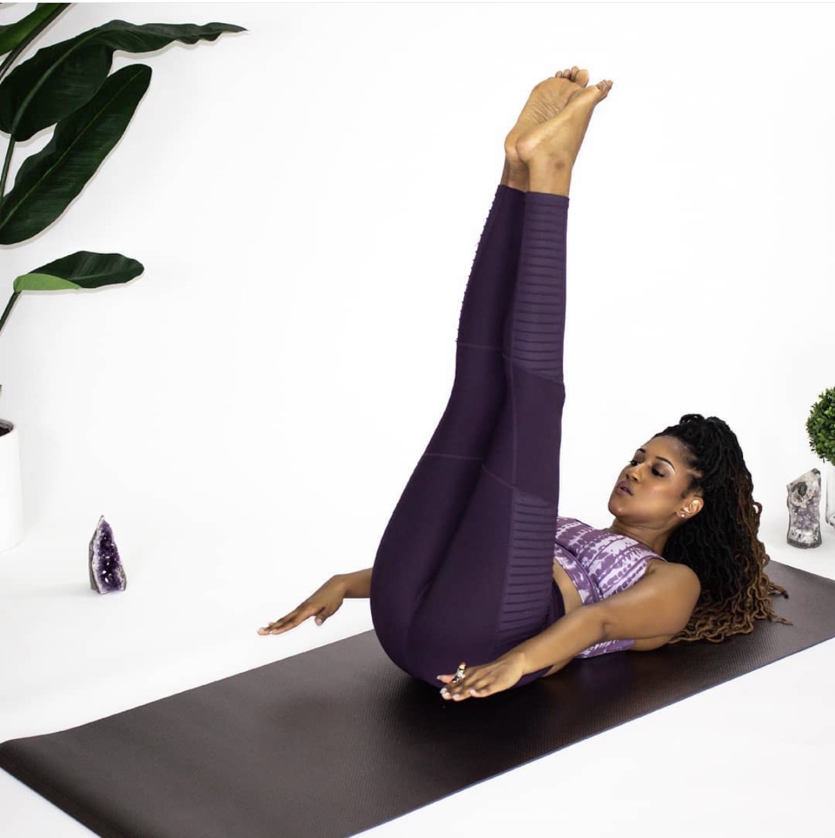Woman practicing yoga on black mat with plants and crystals nearby, lying on her back with legs raised vertically.