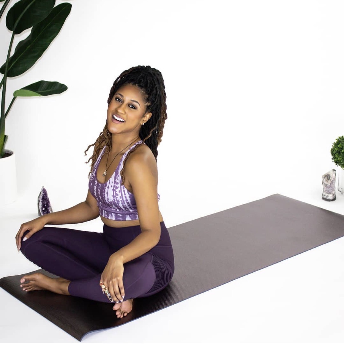 Woman sitting cross-legged on yoga mat, smiling, in a bright room with plants and white walls.