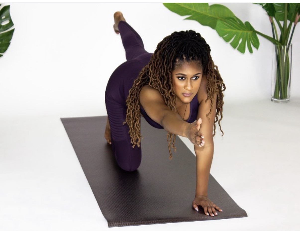 Woman in yoga pose on black mat, reaching forward with one arm, surrounded by green plants, white background.
