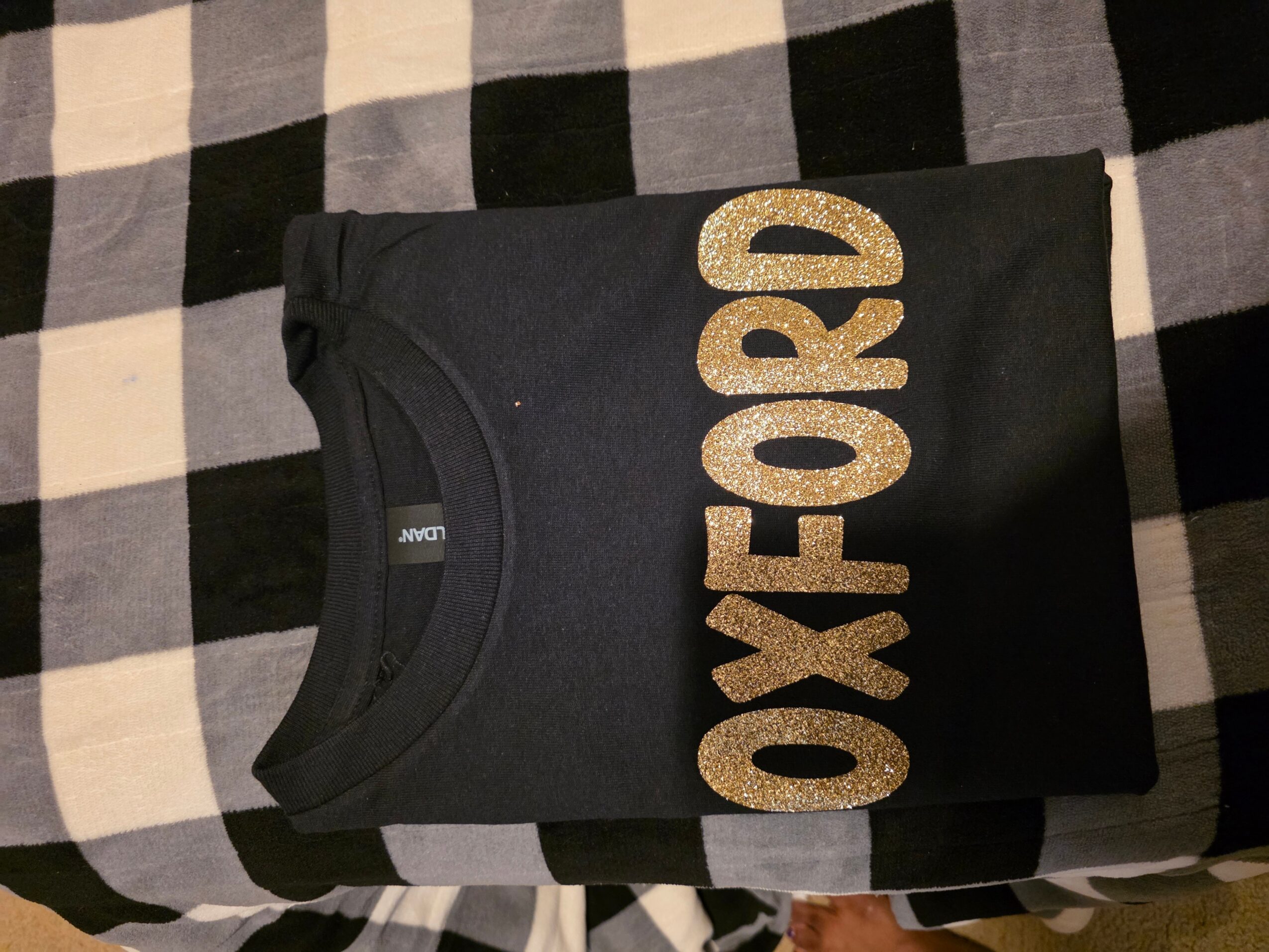 Black T-shirt with large gold glittery text reading 'OXFORD' laid on a checkered fabric surface.