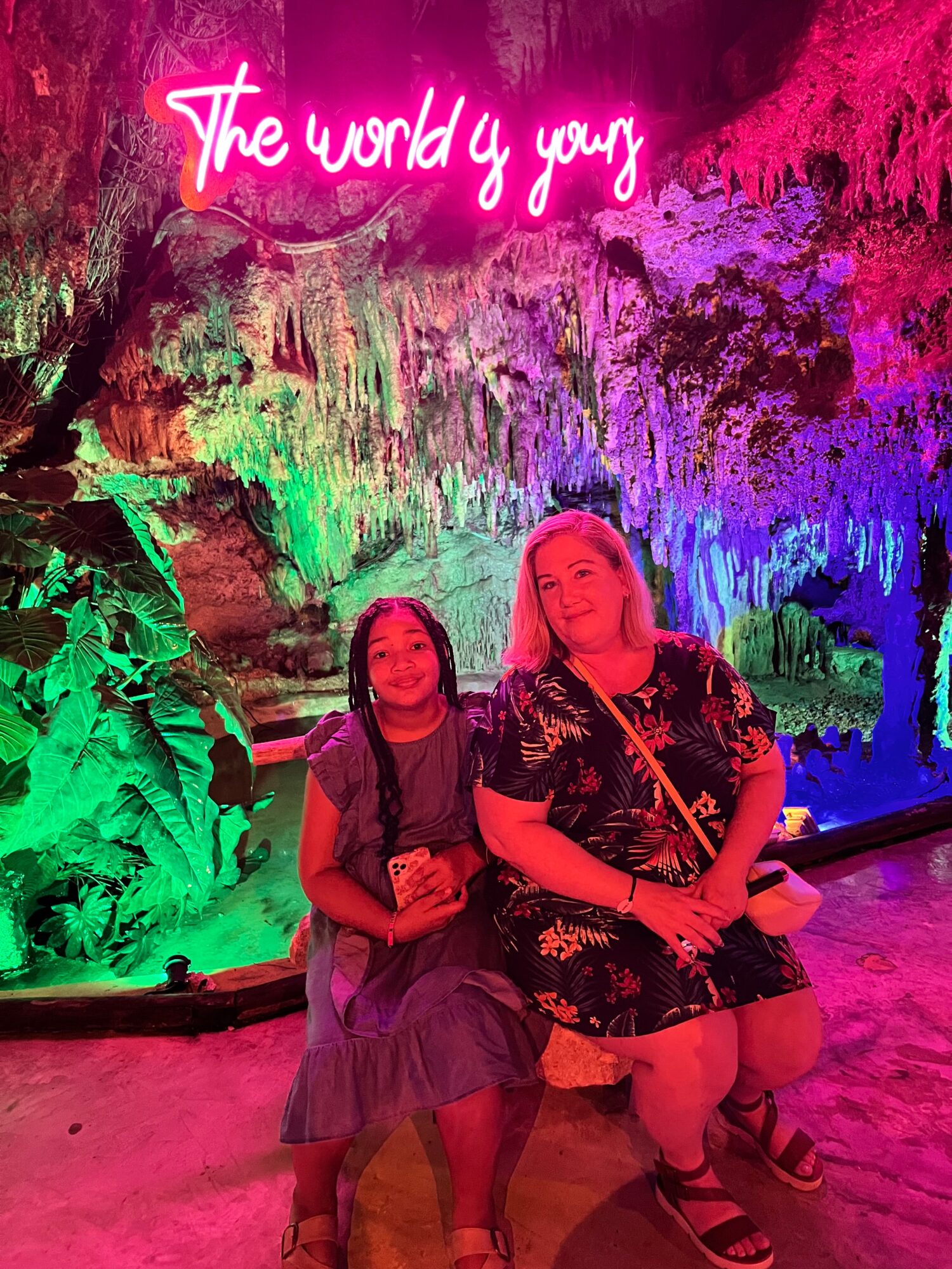 Two women sitting in front of a colorful waterfall display with neon pink text above that reads 'The world y your'.