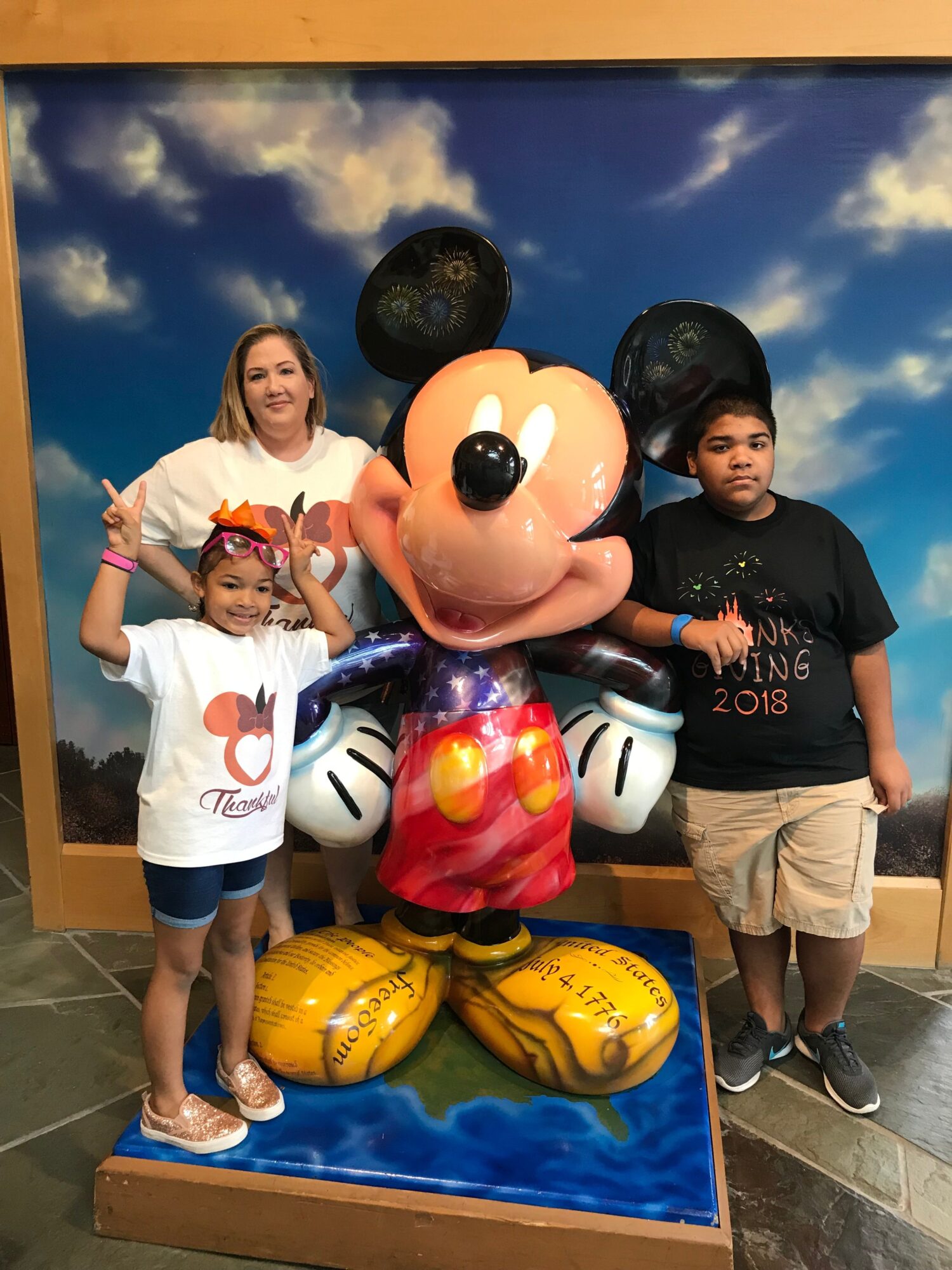 Two children with helmets stand next to a Mickey Mouse statue and yellow footballs on a blue platform.