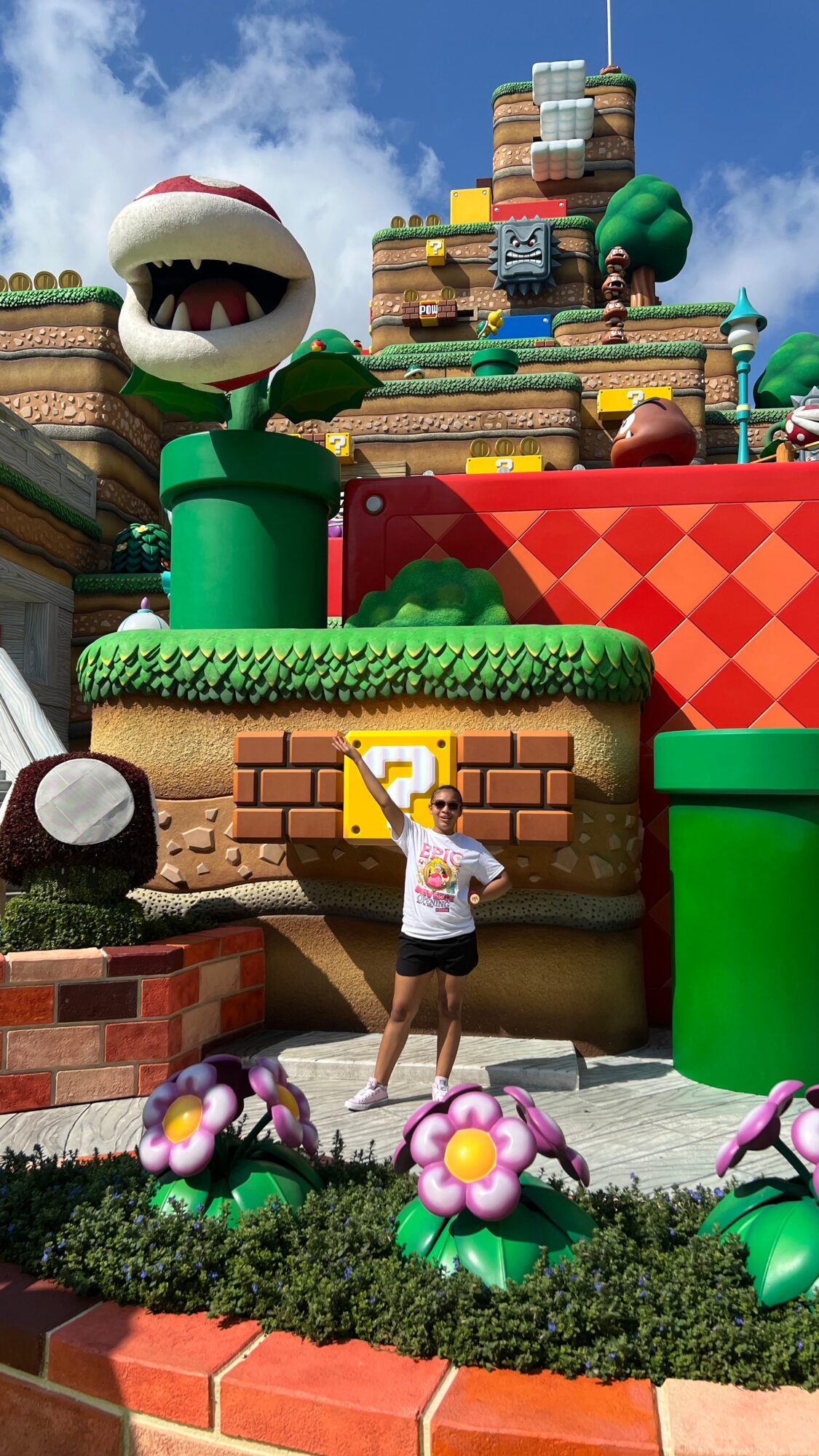 Person standing in front of a large Super Mario-themed display with a giant Bowser head and colorful stacked blocks, flowers in foreground.