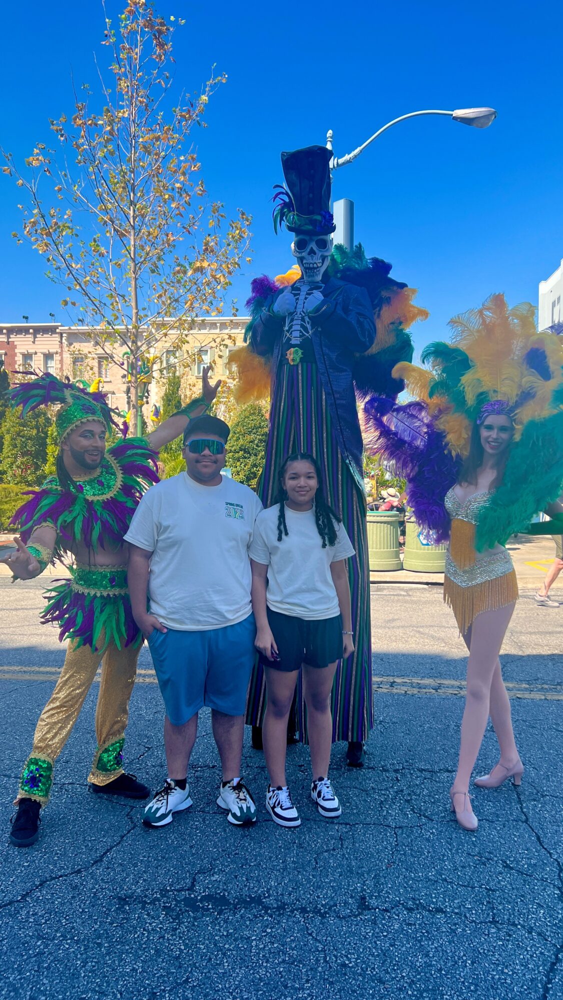 Two children stand with three performers in colorful costumes and a tall puppet on a city street.