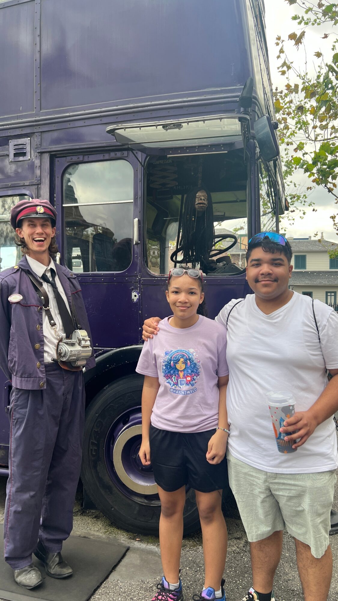 Three people standing in front of a large purple vehicle, smiling, with trees and a building in the background.
