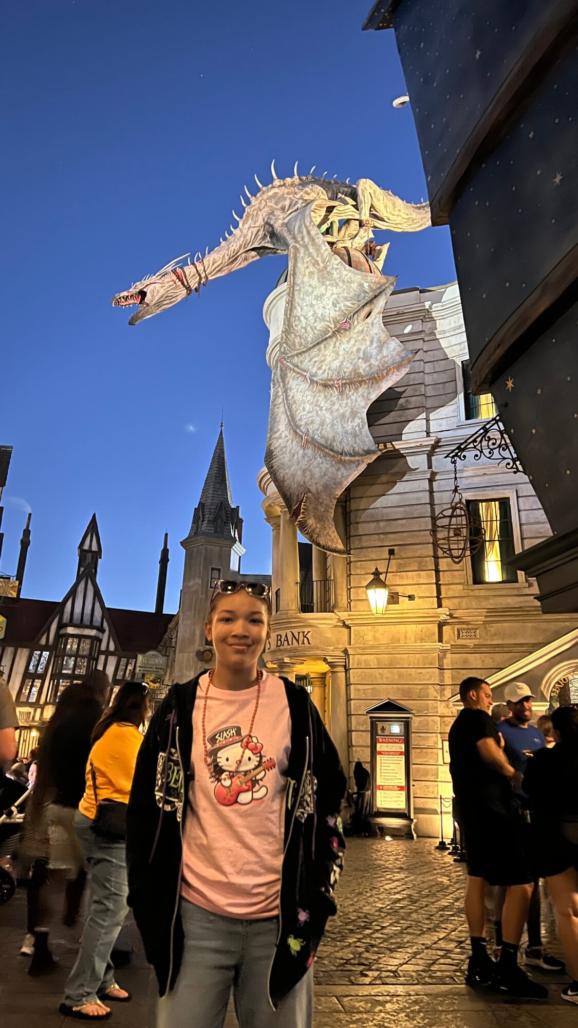 A person in a pink shirt and black jacket stands in a themed outdoor area with buildings and a dragon hanging overhead against a blue sky.