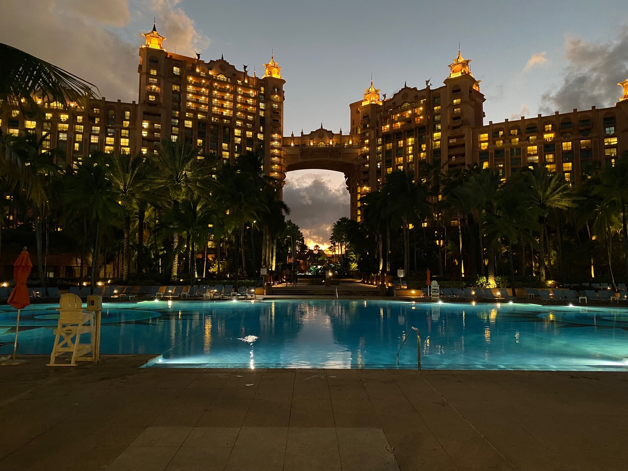 Luxury hotel with towers and an archway, illuminated at dusk, reflected in a swimming pool surrounded by palm trees.