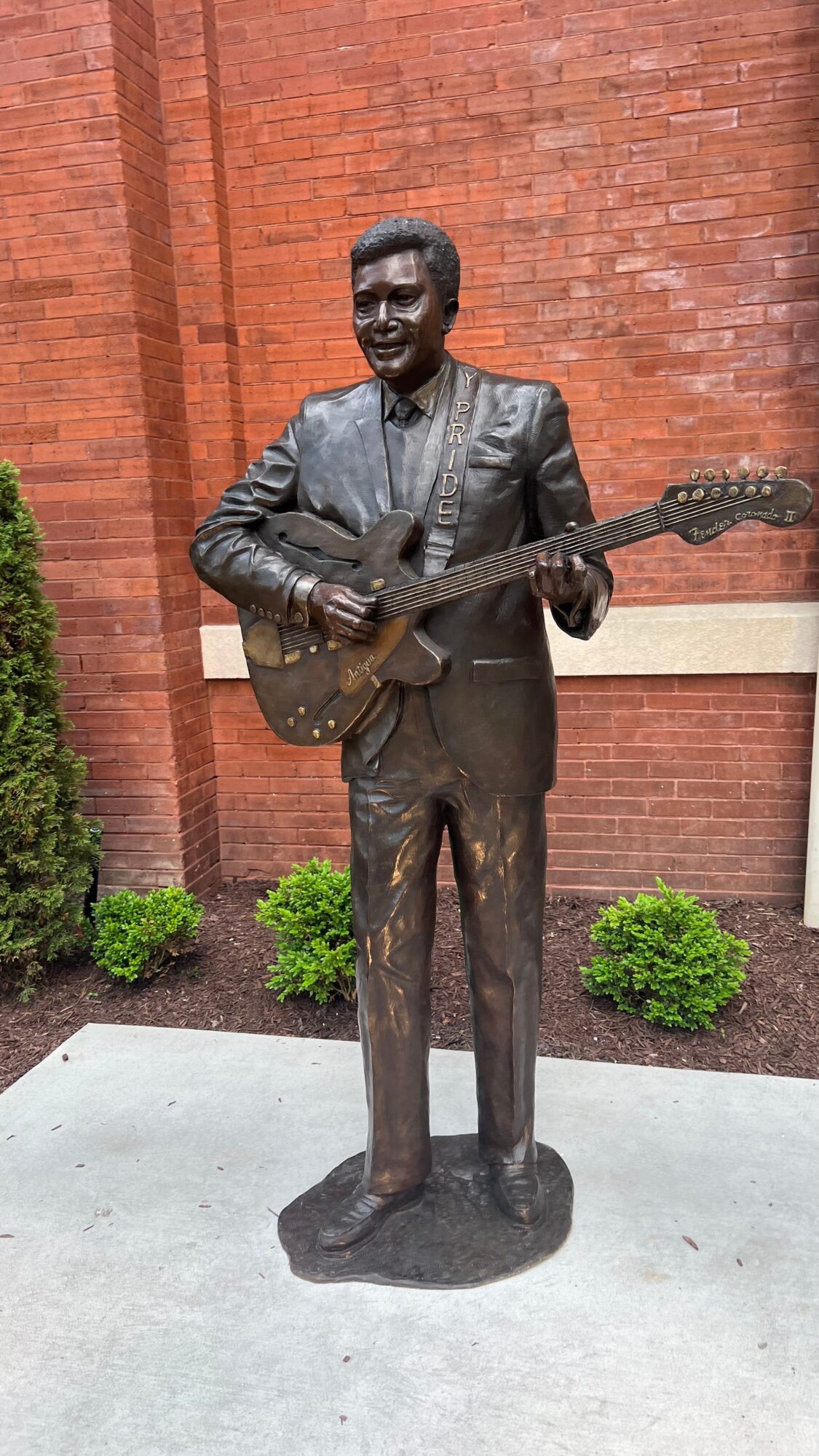 Bronze statue of a man in a suit playing an electric guitar, standing outdoors in front of a brick wall.