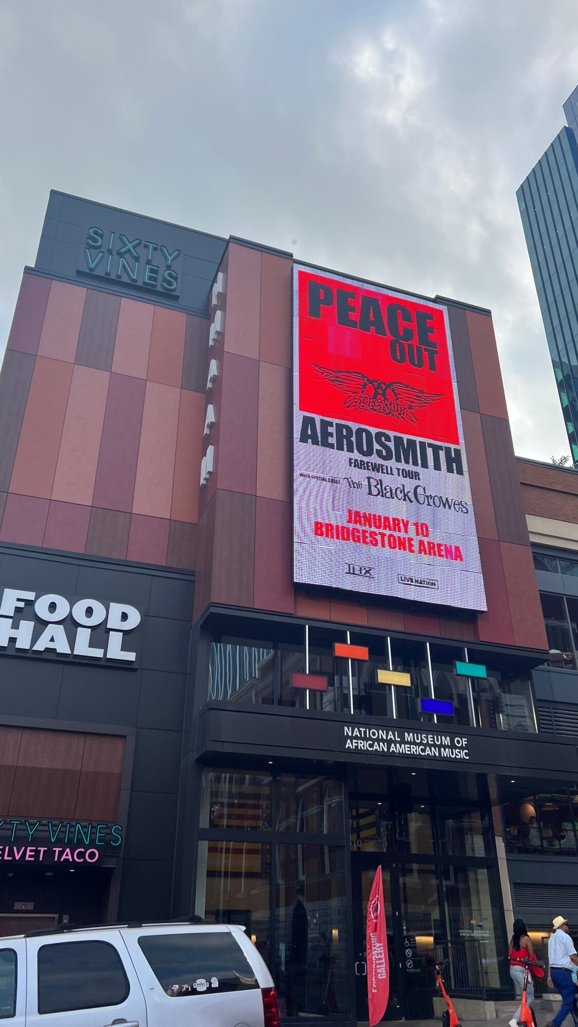Large digital billboard displays concert information, with a building and cloudy sky in the background.