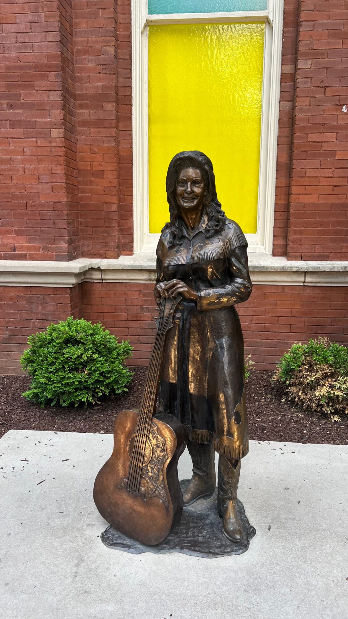 Bronze statue of a woman holding a bag, standing in front of a brick building with a yellow window, surrounded by bushes.
