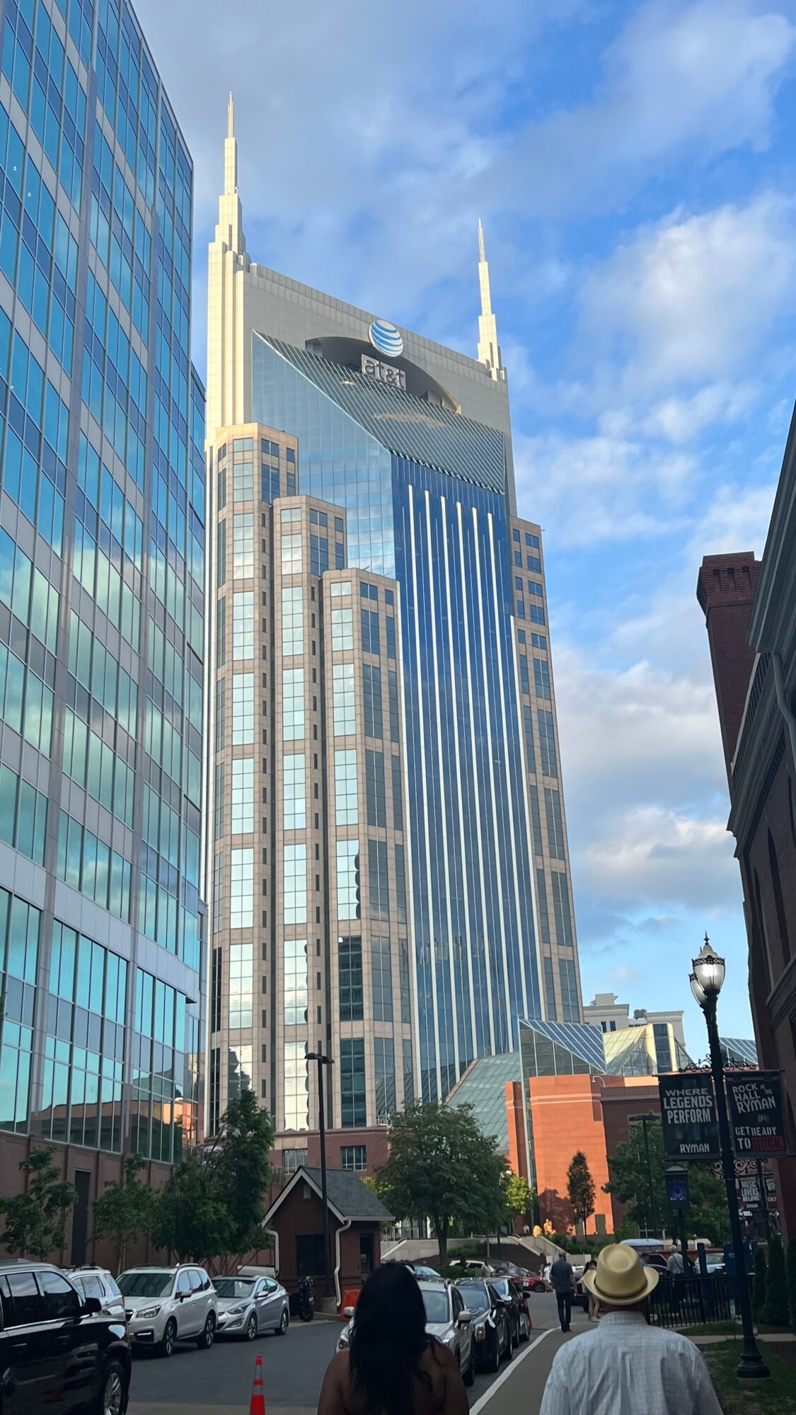 Tall skyscraper with spires and a digital billboard, surrounded by other buildings and a street scene with cars and pedestrians.