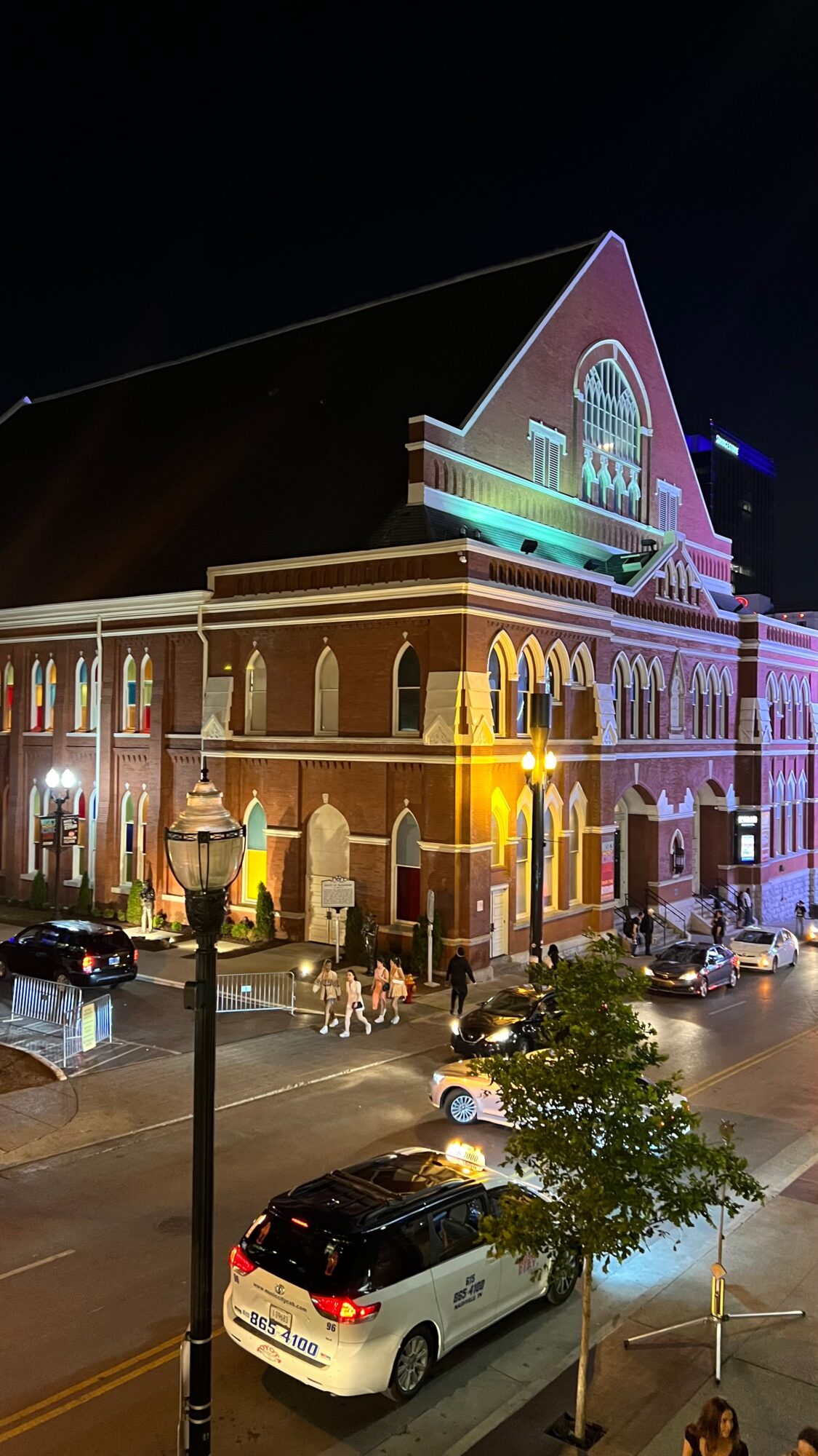 Night view of a large brick building with colorful lights, cars parked in front, and a tree on the street.