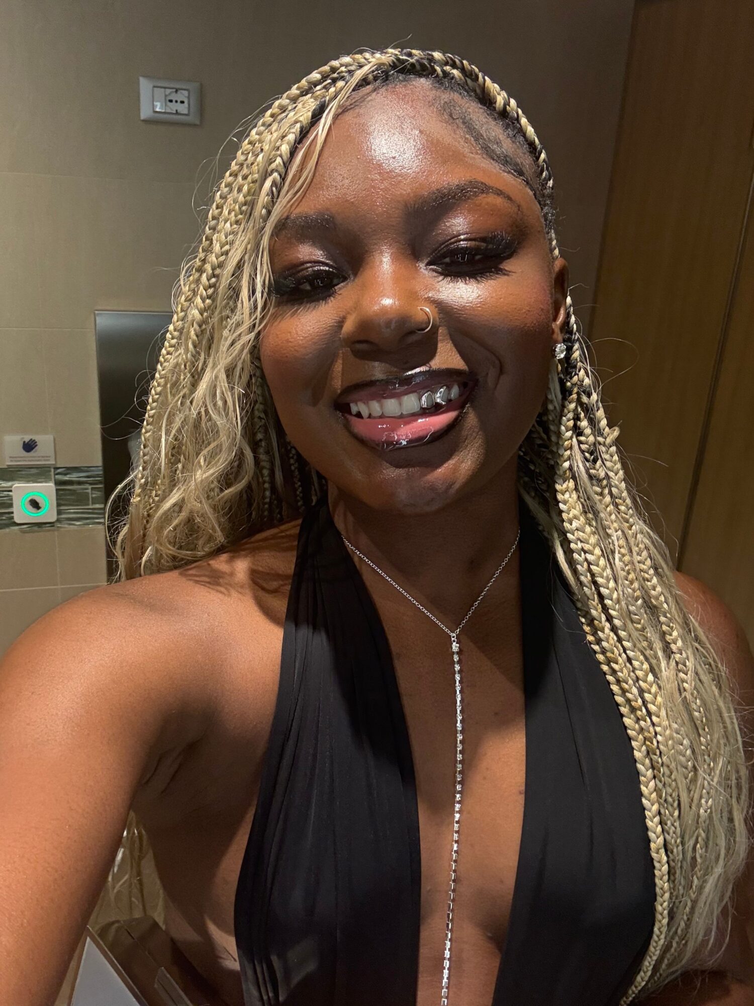 Smiling woman with braided blonde hair, wearing a black dress and pearl necklace, taking a selfie indoors.