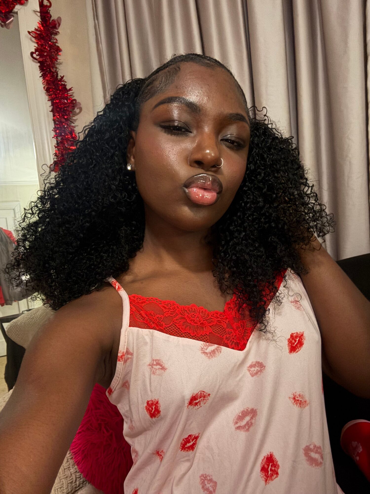 Young woman with curly hair wearing a white dress with red accents, lying on a bed with curtains in the background.