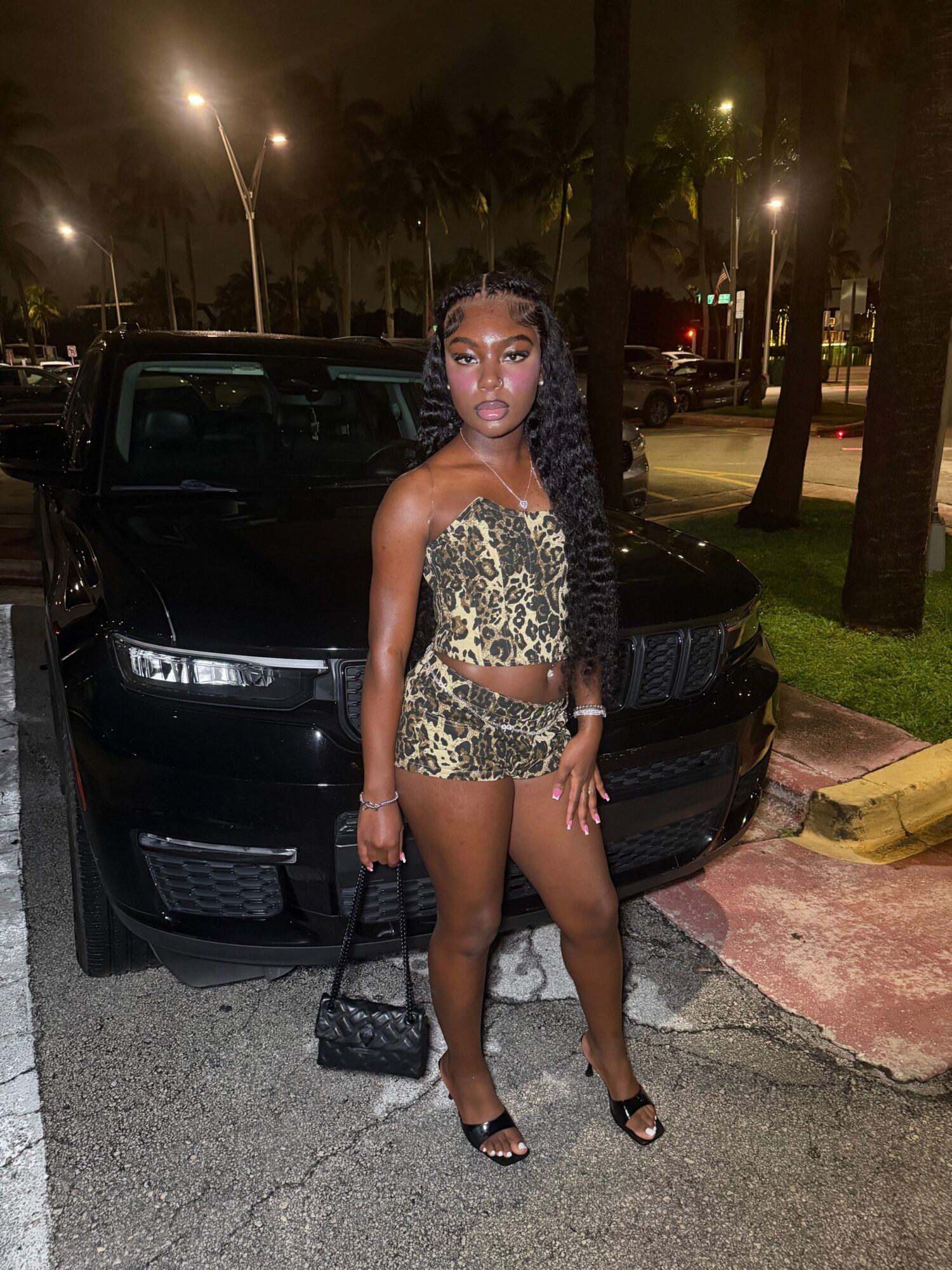 Woman in animal print outfit standing next to a black car at night, illuminated by streetlights.