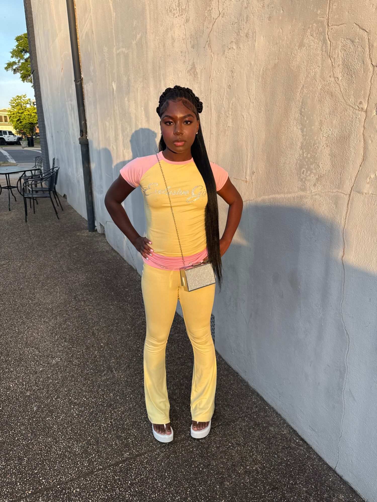 Young woman with braided hair standing outdoors against a wall, wearing yellow pants and a pink top, carrying a bag.