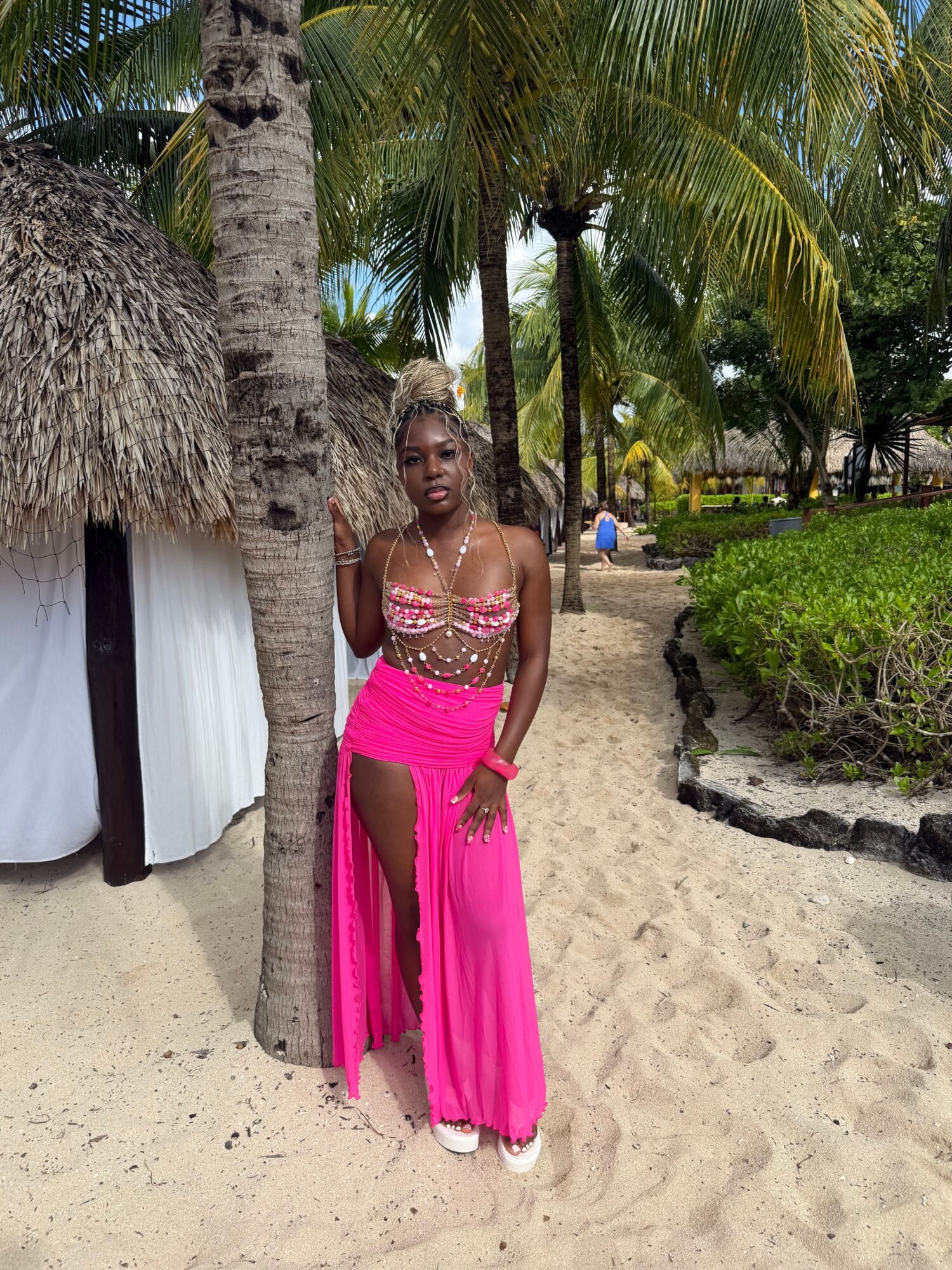 Woman in pink dress leaning against a palm tree on a sandy beach with greenery in the background.