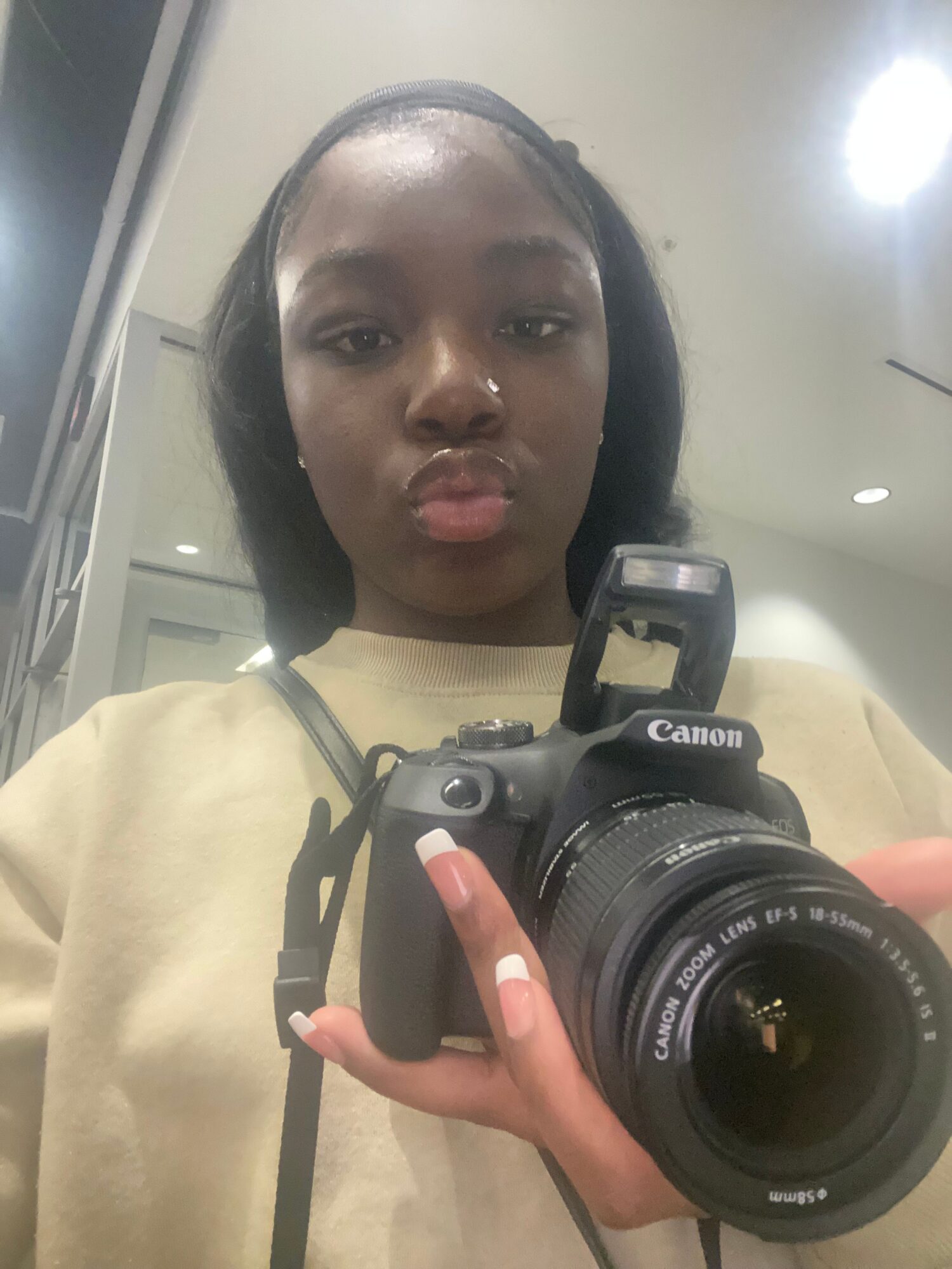 Person taking a mirror selfie with a camera, face visible, holding the camera with one hand, background blurred.