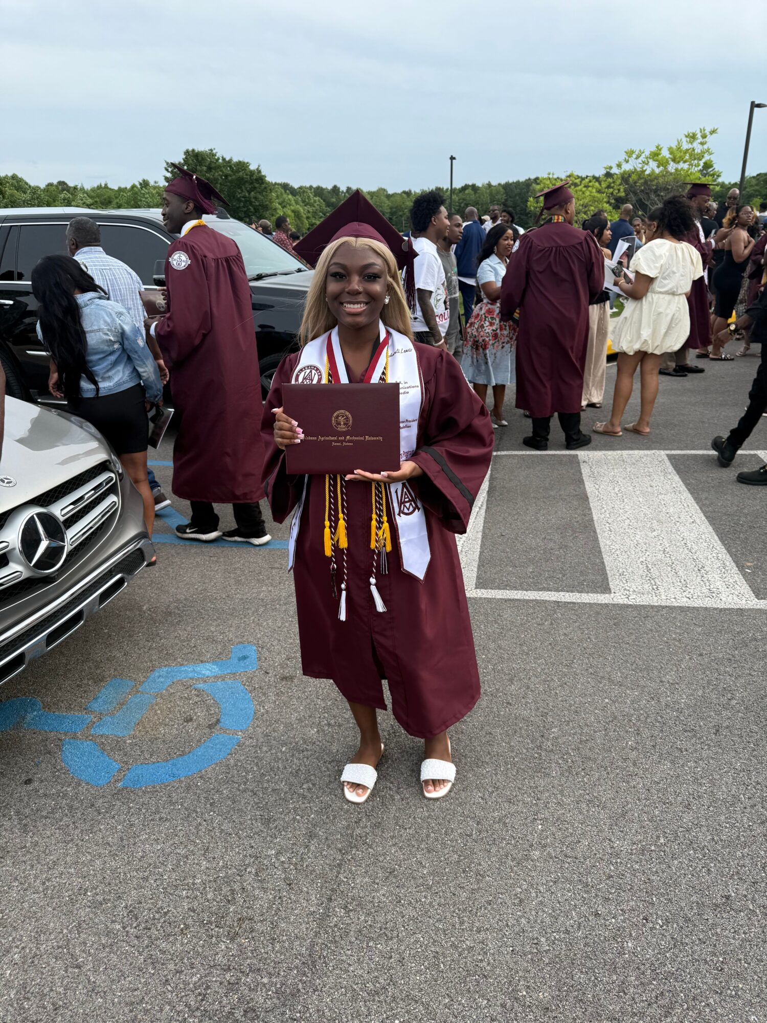 Person in graduation gown and cap standing on a parking lot, holding a diploma, with other people in the background.
