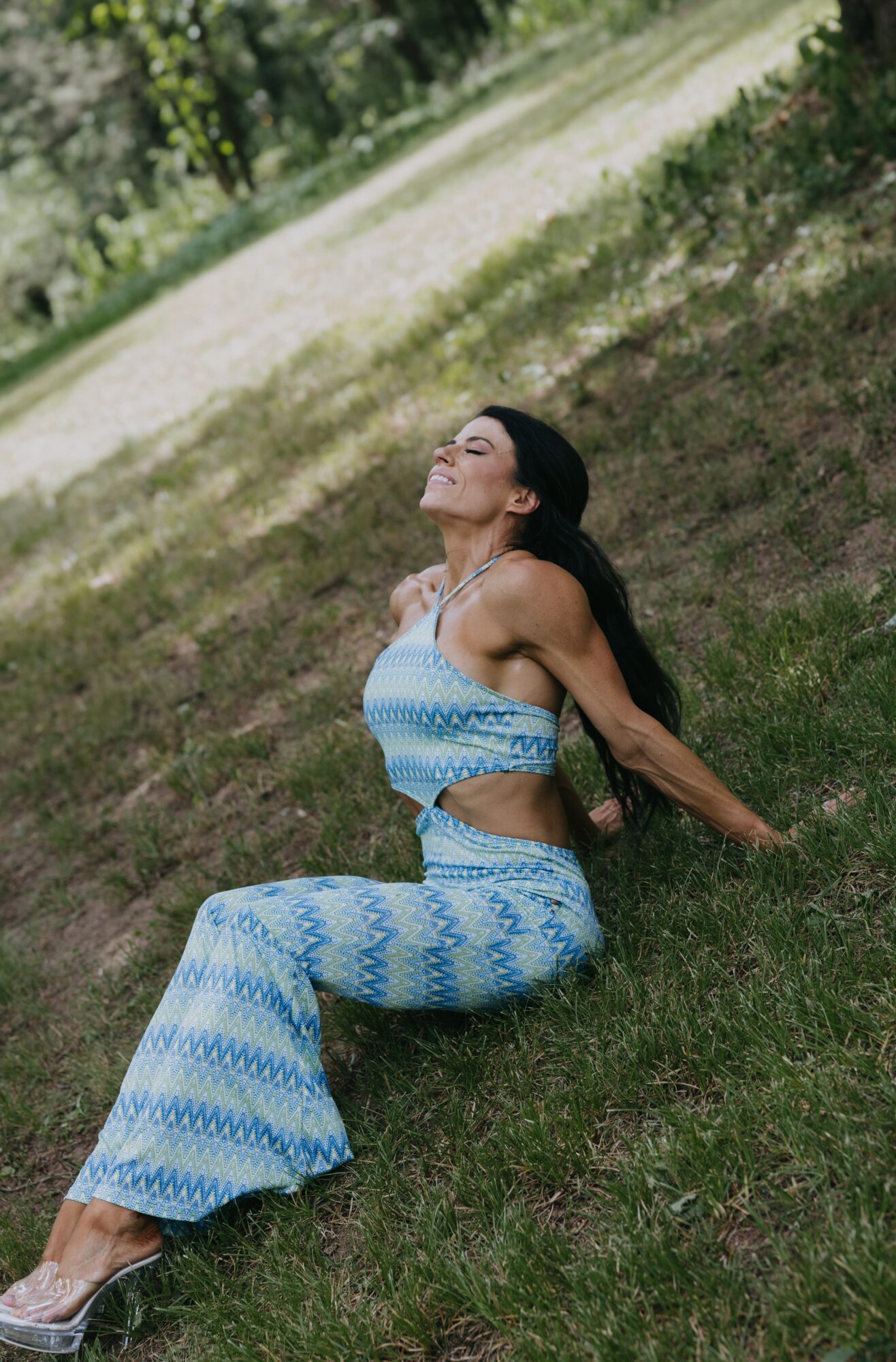 Woman sitting on grass with eyes closed, wearing a blue and white patterned outfit, outdoors in a park-like setting.