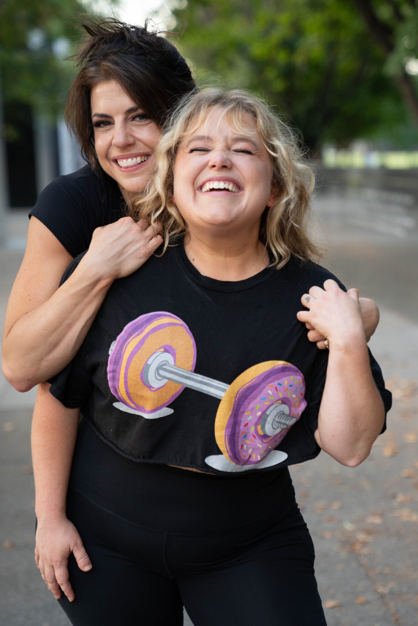 Two women smiling outdoors, one holding a colorful toy dumbbell, surrounded by trees and a paved path.
