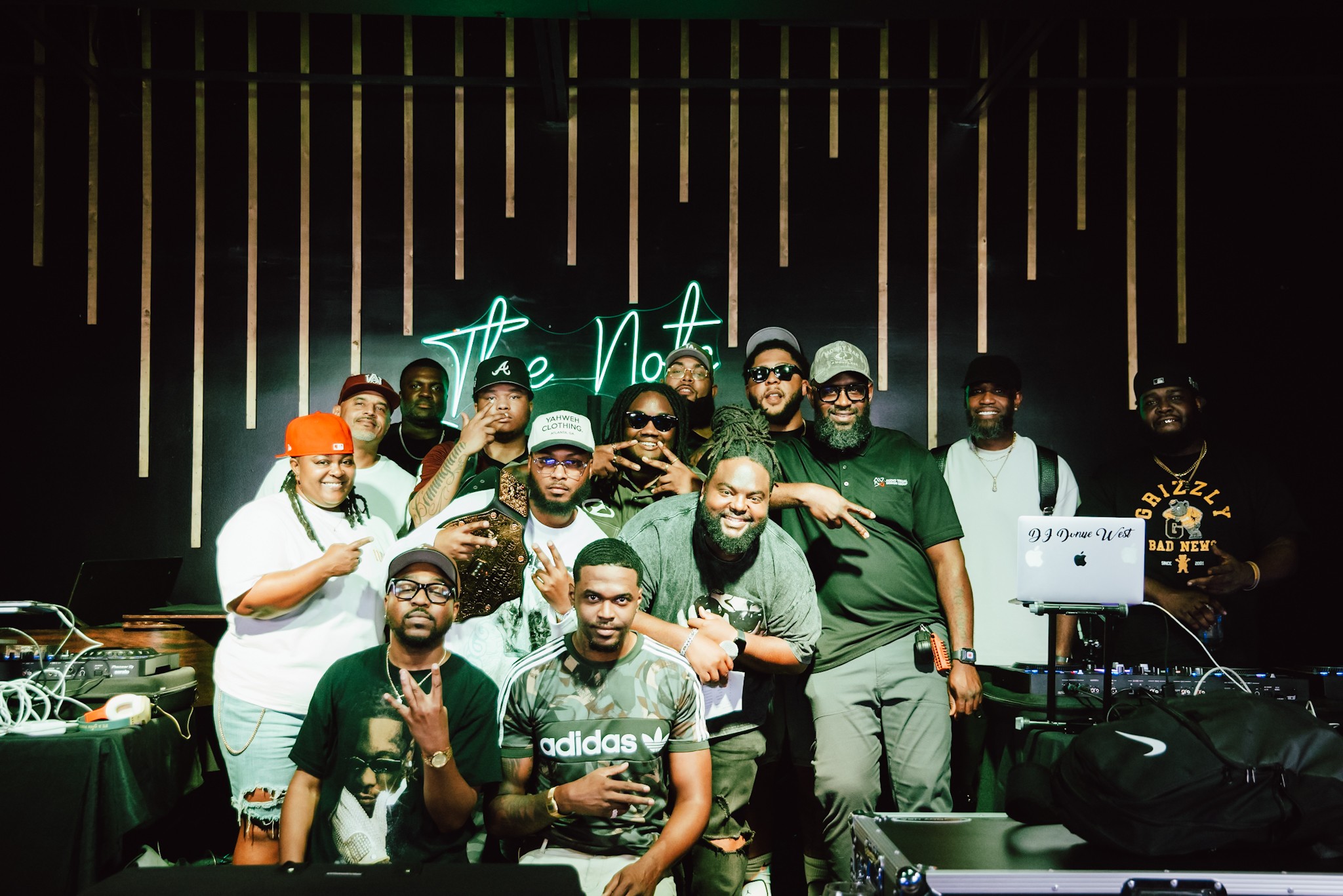 Group of people on stage with DJ equipment, some making peace signs, in front of a backdrop with vertical gold lines.
