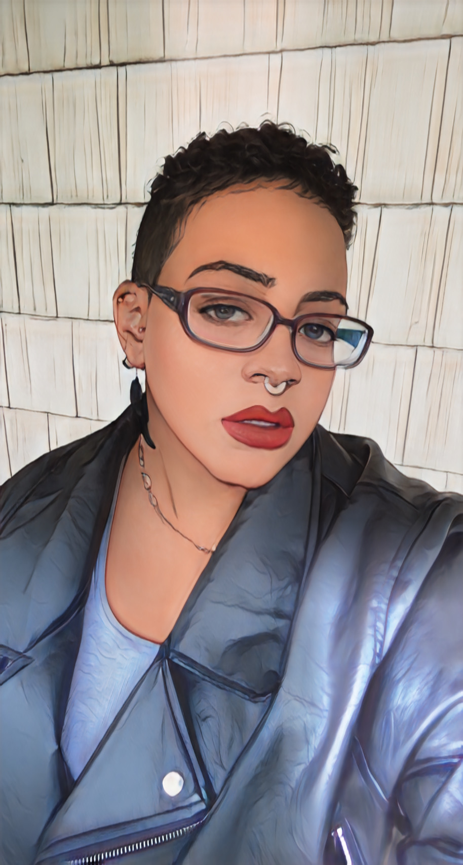 Person with short curly hair, glasses, red lipstick, and a septum piercing, wearing a leather jacket and a necklace, against a tiled background.