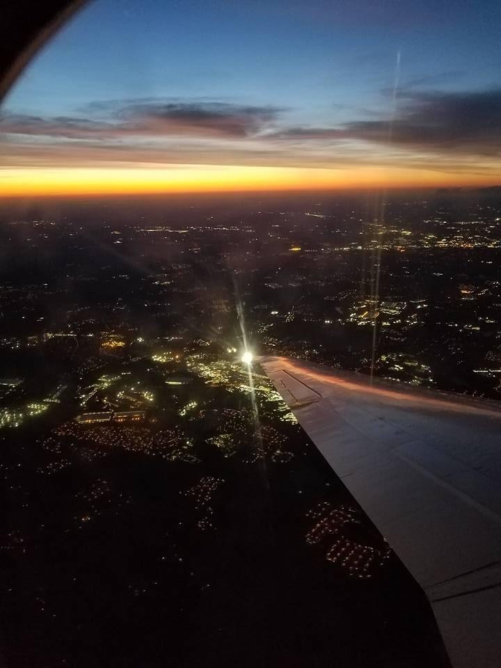 A view from an airplane window showing a cityscape at dusk with a colorful sunset sky and city lights below.