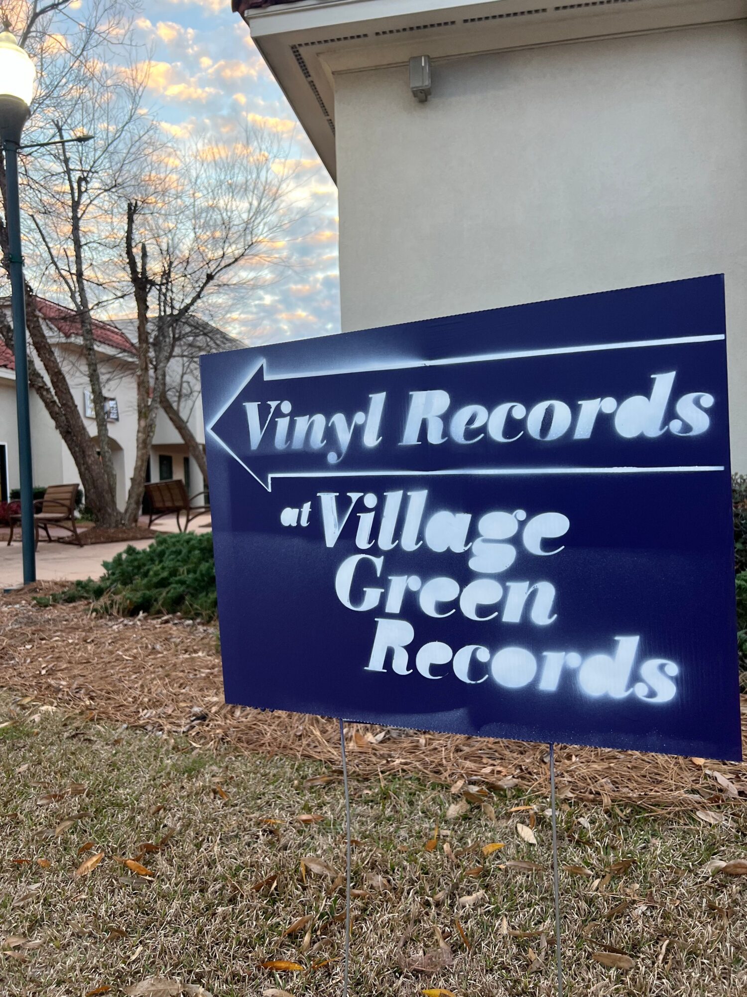 Sign for Village Green Records with white text on a blue background, outdoors with trees and ground visible.