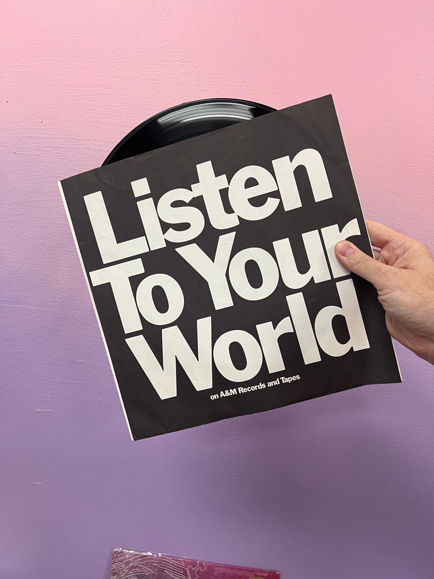 Hand holding a black sign with white text reading 'Listen to your World' against a pink and purple background.