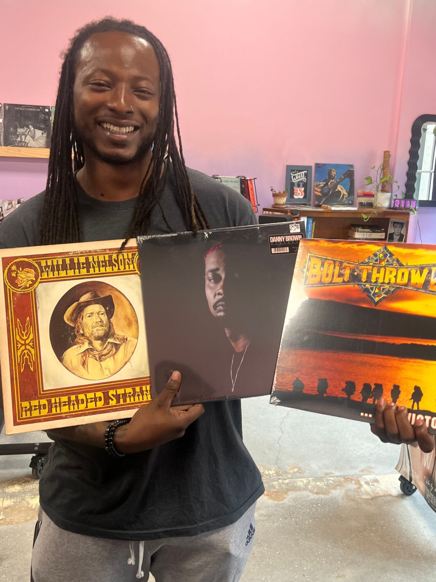 Person holding a vinyl record cover with a woman and a framed portrait of a woman, in a room with pink walls and posters.