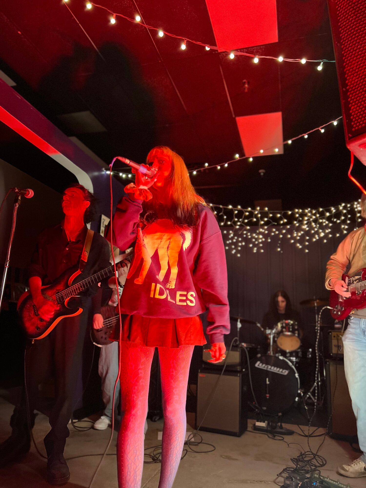 Band performing on stage with string lights overhead, person with red hair singing, others playing instruments, dim lighting.