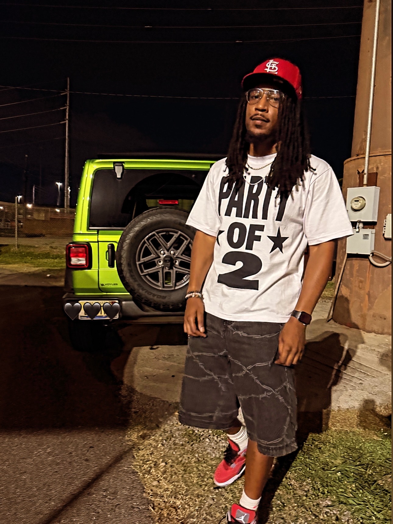 Young man with long dreadlocks wearing a white T-shirt, black shorts, red sneakers, and a red cap, standing outside at night.
