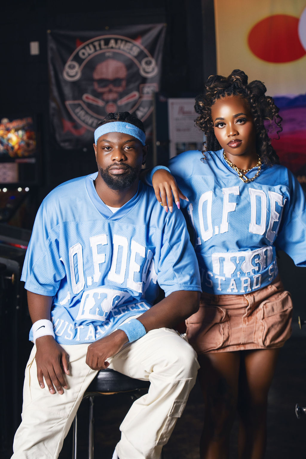 A man and woman pose together, both wearing blue shirts with white text, in a dimly lit indoor setting.