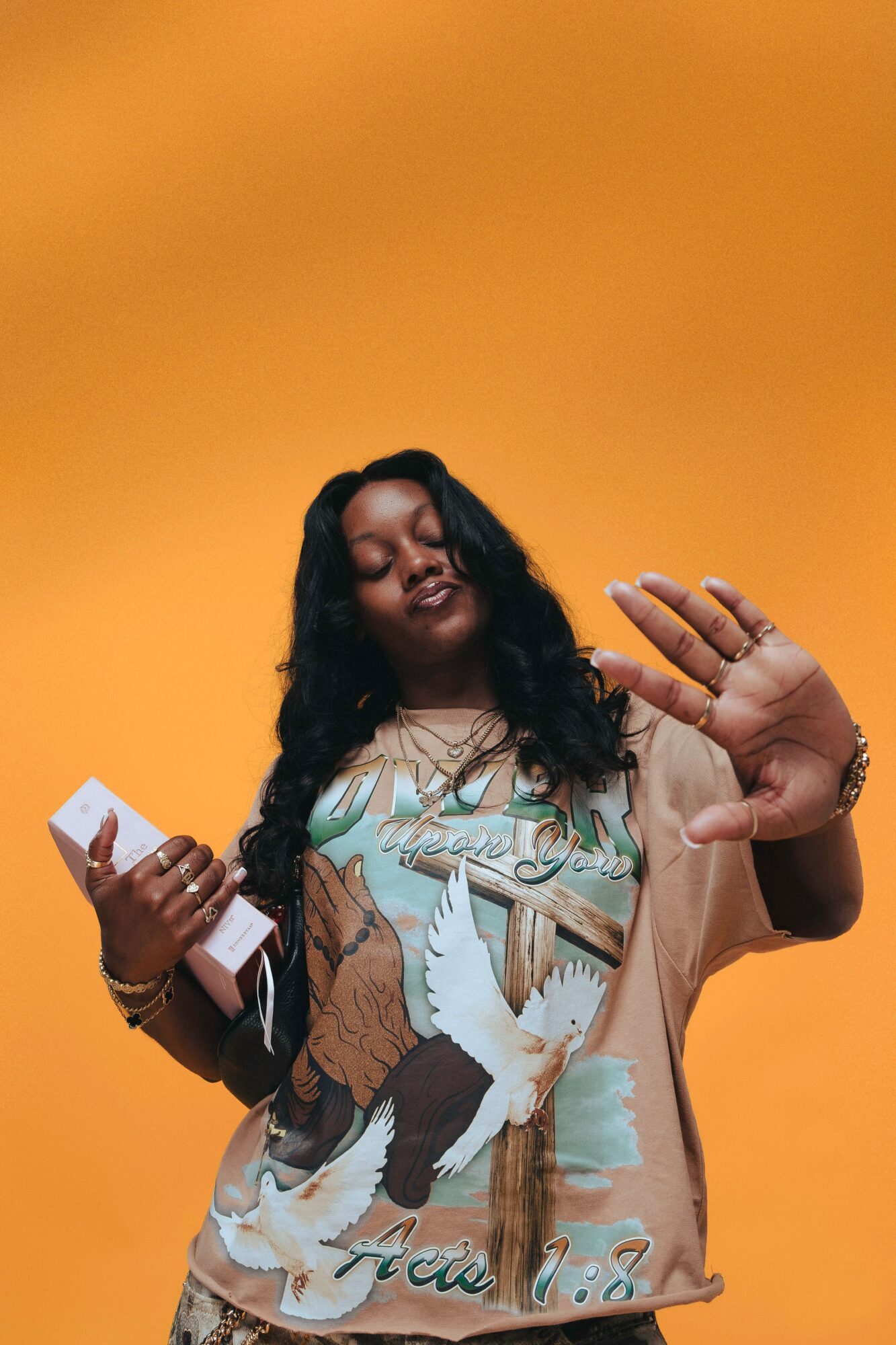 Woman with long dark hair holding a phone and raising her hand against an orange background.