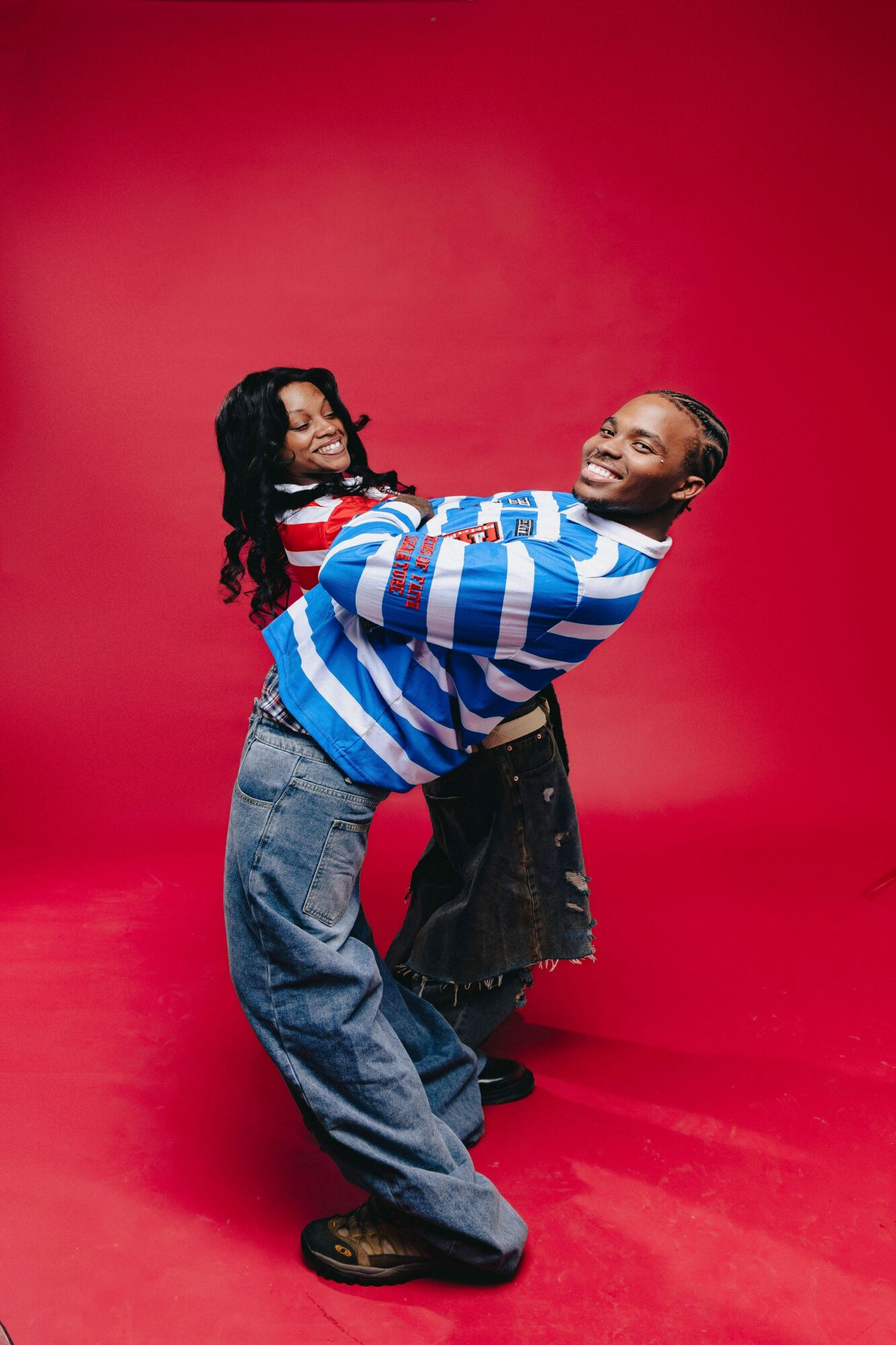 Two people dancing against a red background, smiling and holding each other, with casual clothing and relaxed poses.