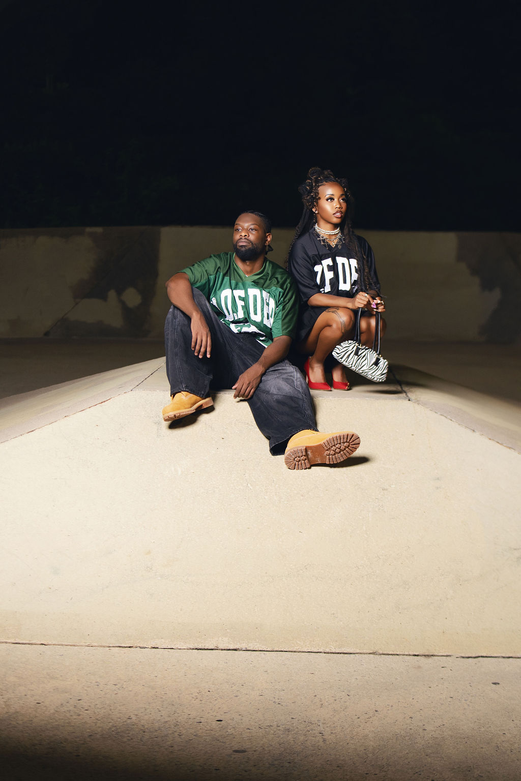Two people sit on a concrete surface at night, one with a beard and the other with braided hair, both wearing black and green shirts.