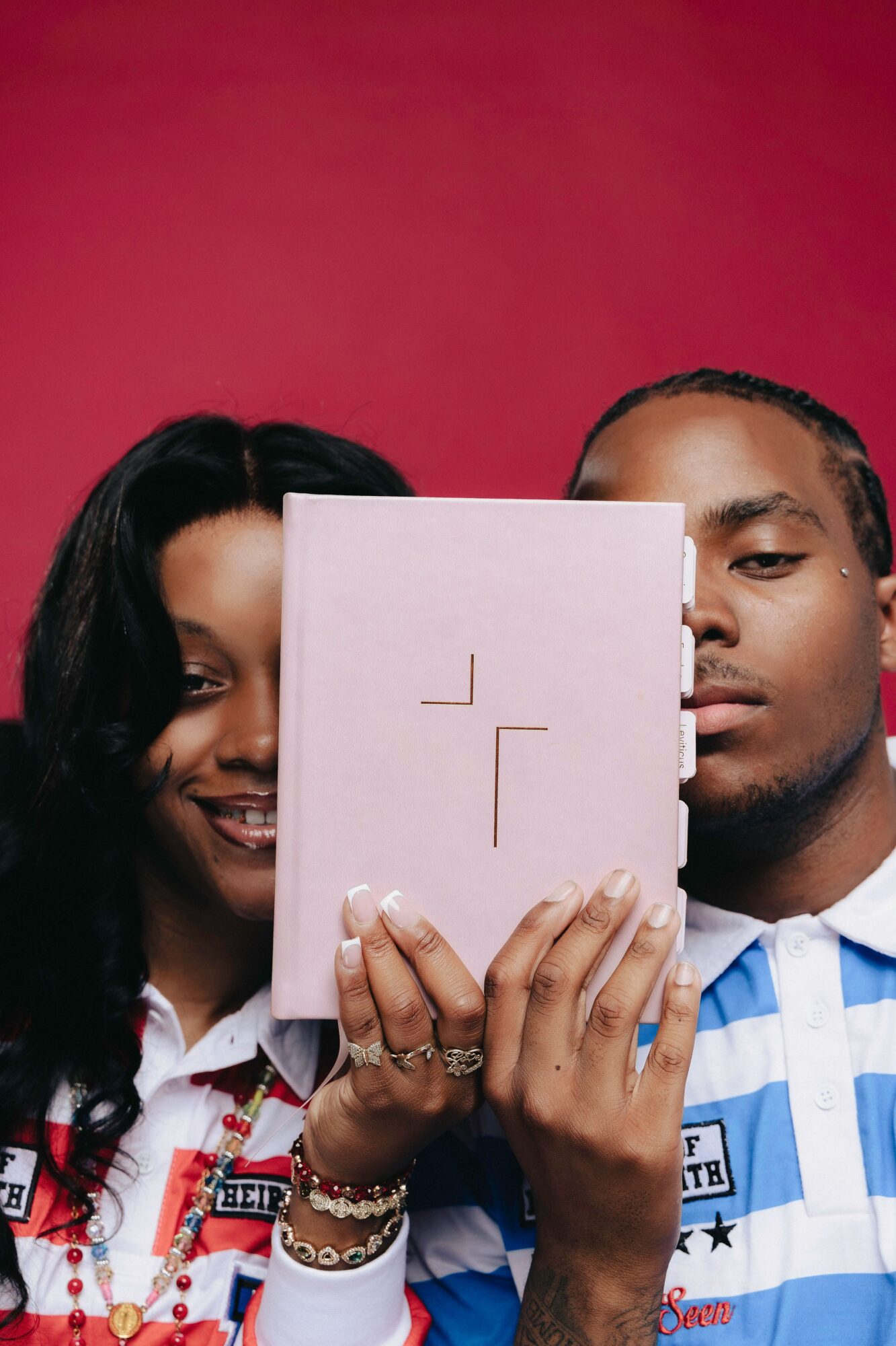 Two people holding a pink book in front of their faces, with a red background, one smiling and one serious, wearing striped shirts.
