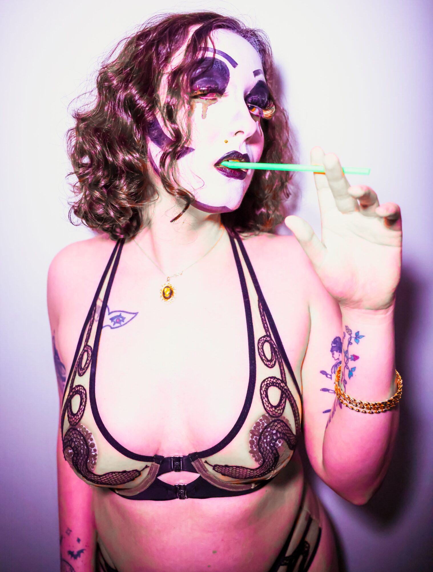 Person with curly hair and makeup, wearing a lace top, holding a green straw, with tattoos and jewelry, against a plain background.