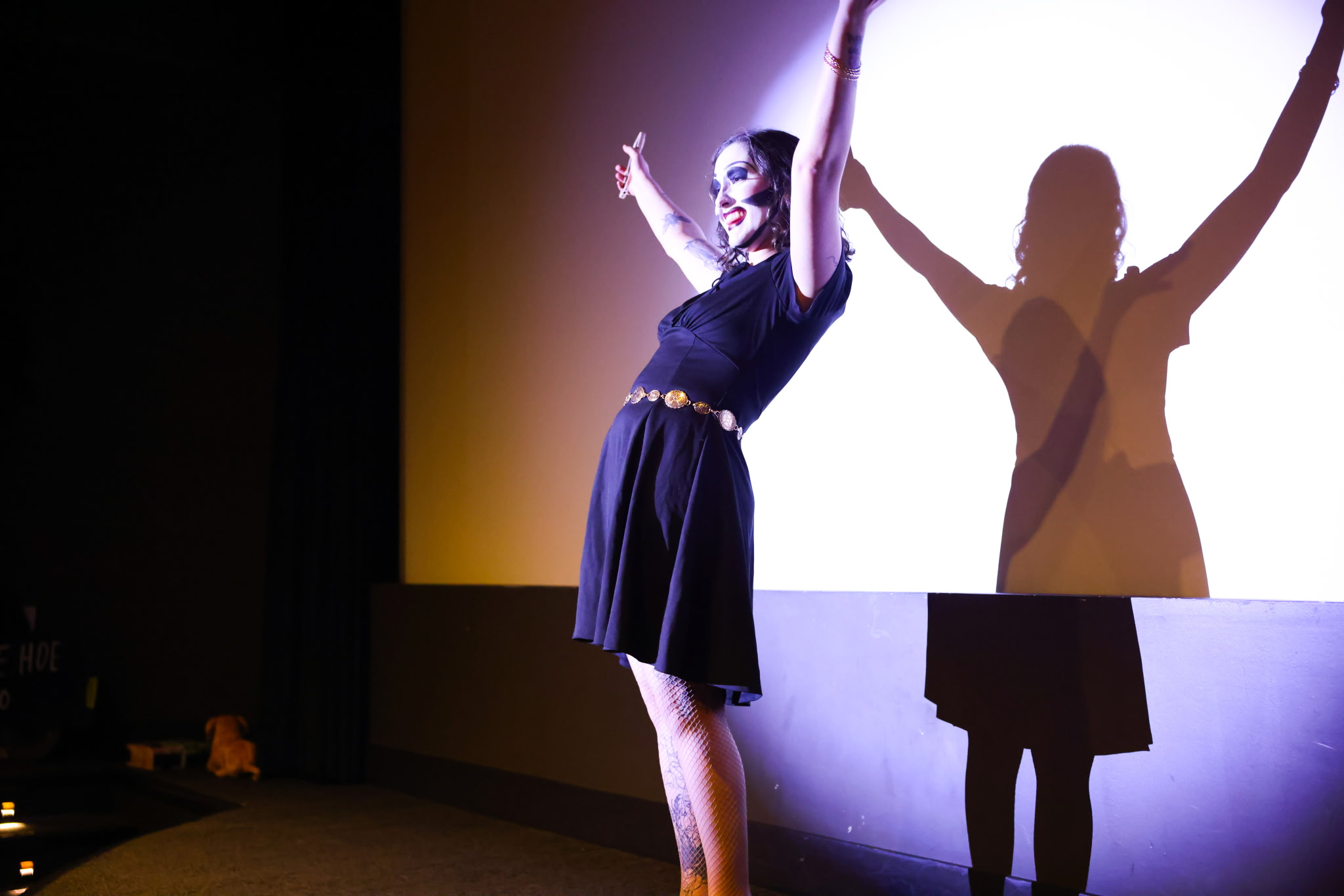 Performer on stage with arms raised, casting a shadow on a bright screen behind her, wearing a dark dress with a belt.