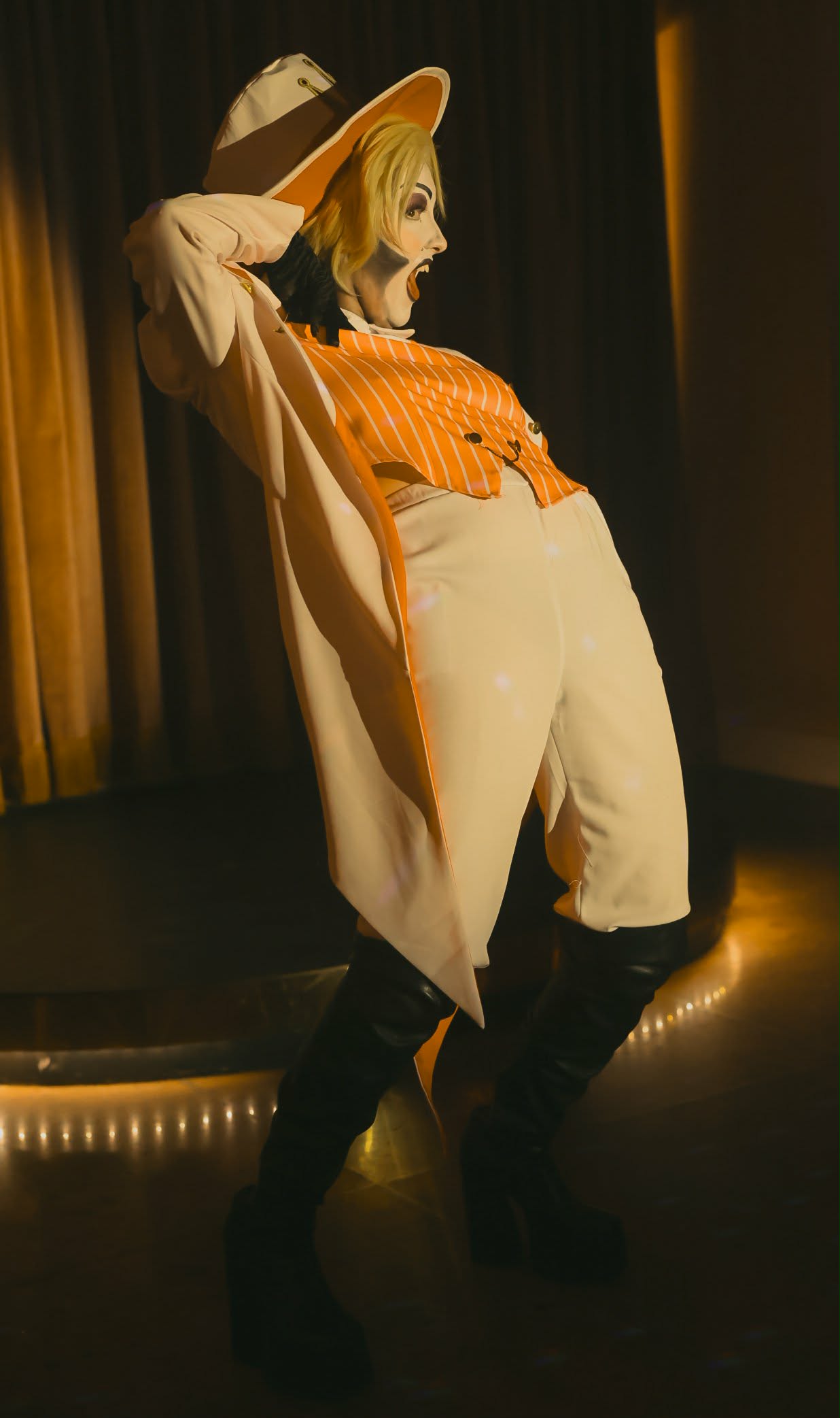 Person in orange top, white pants, black boots, and hat, posing on stage with curtains in background.