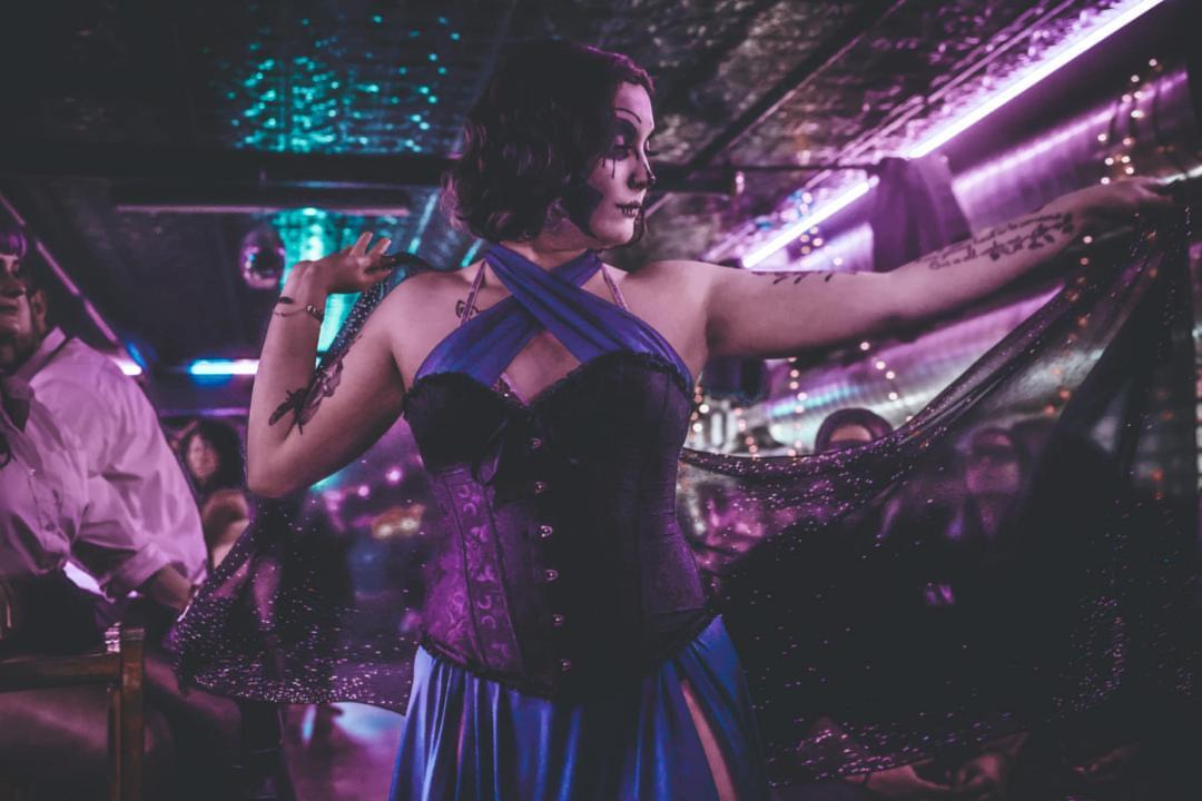 Woman with short hair and makeup in a colorful, illuminated nightclub, wearing a dark, strapless dress with blue accents, dancing.