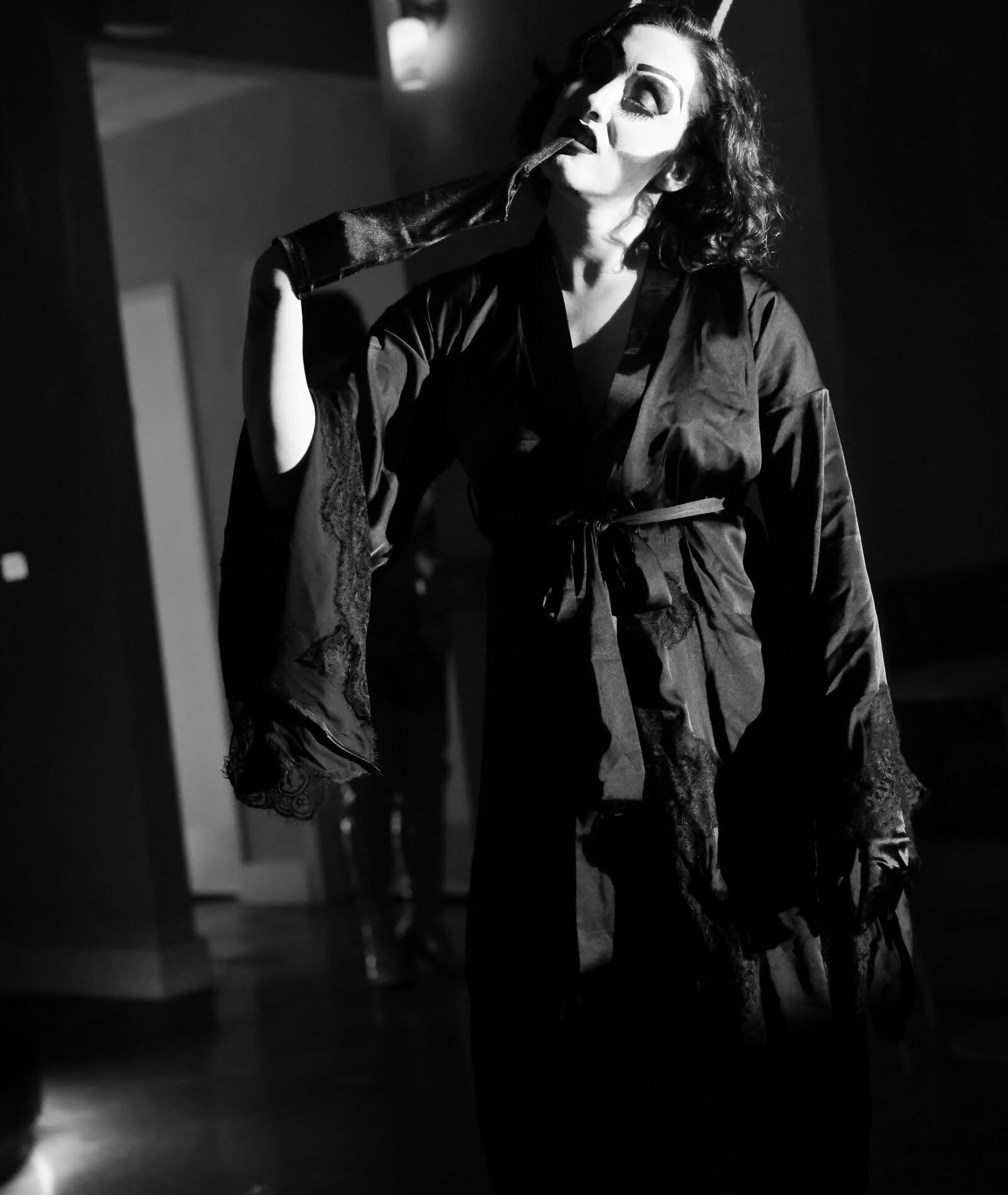 Person in costume with makeup, wearing dark clothing, holding a mask near face, in a dimly lit room.
